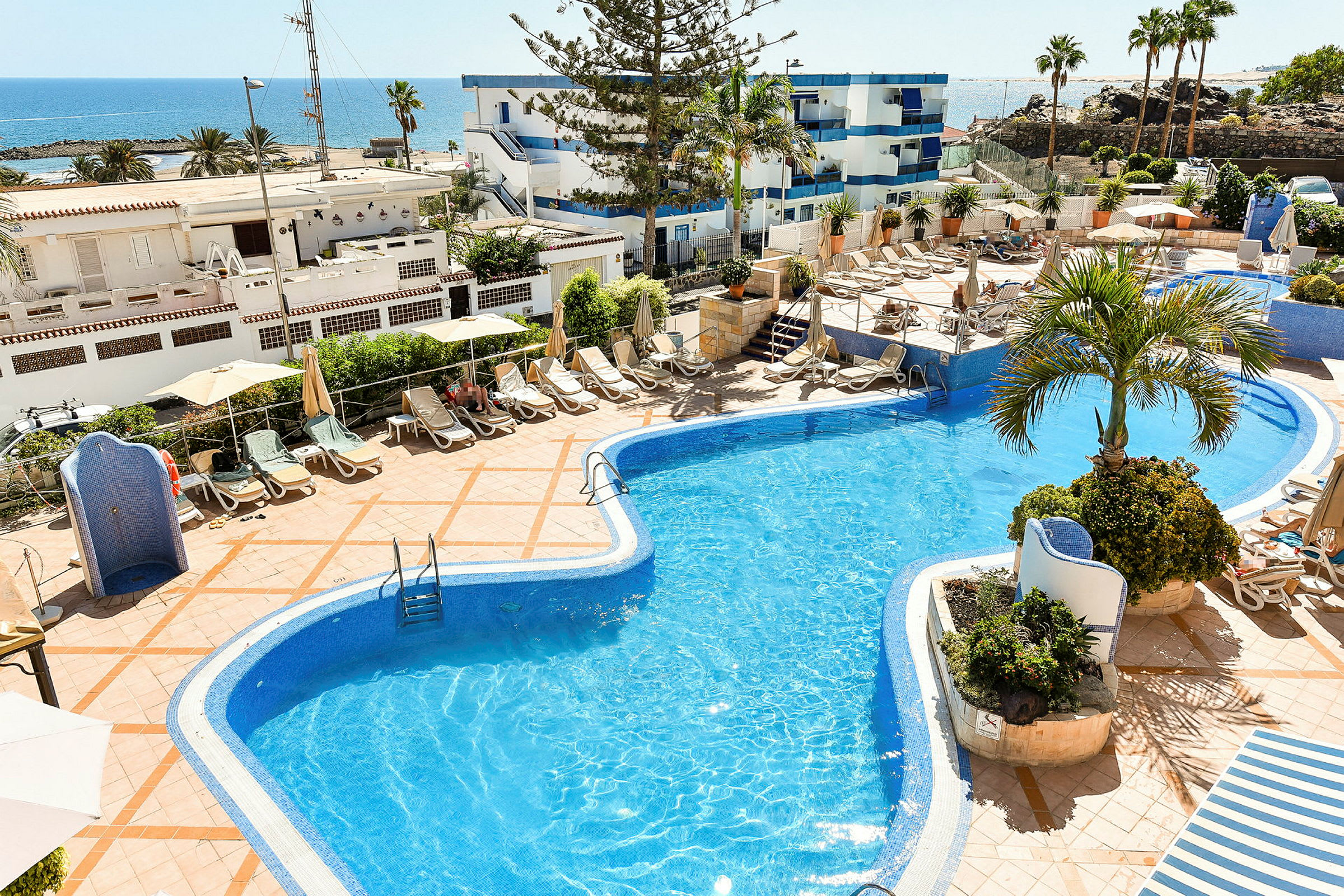 En pool med liggestole og parasoller på omkringliggende terrasse. I baggrunden ses havet og bygninger. (AI-genereret tekst)