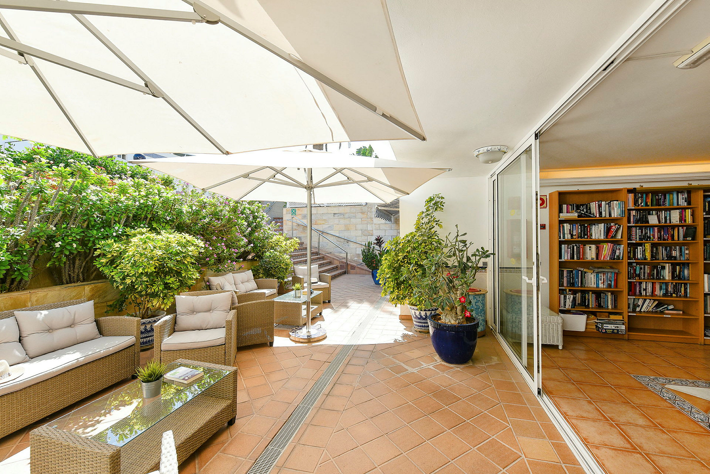 Havemøbler og parasoller på en terrasse med beplantning, der støder op til et rum med bogreoler. (AI-genereret tekst)