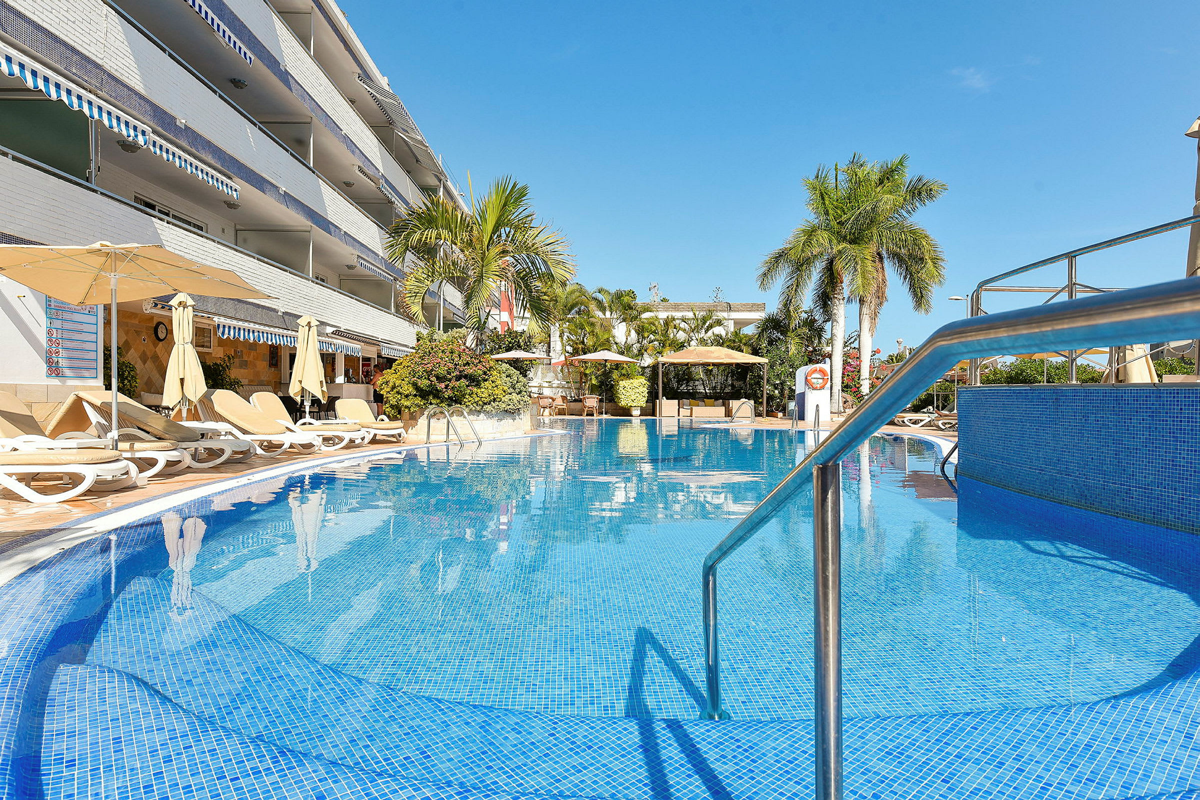Udendørs swimmingpool ved hotel med liggestole og parasoller, omgivet af palmer. (AI-genereret tekst)