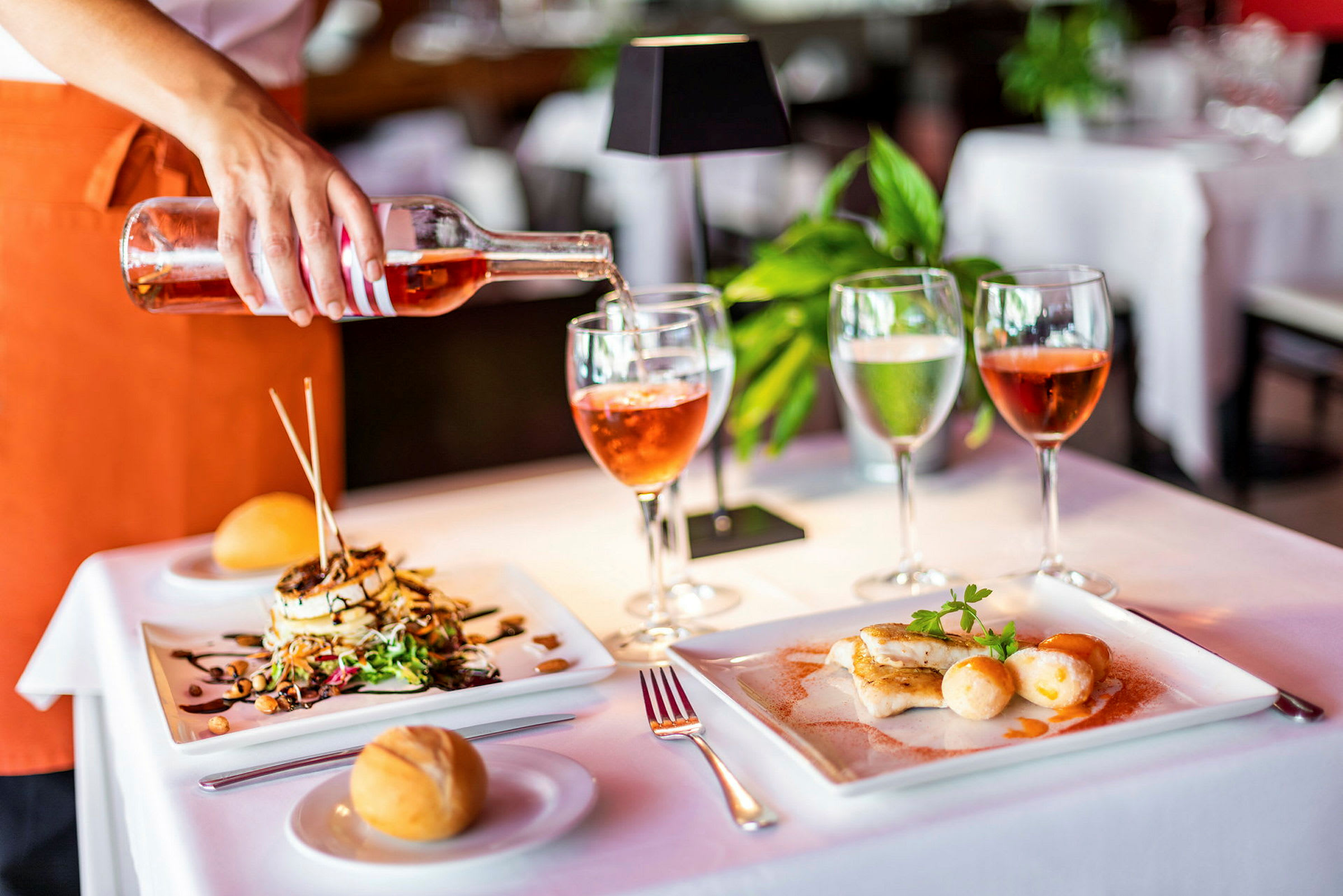 Et bord dækket med tallerkener med mad, glas og brød, samt en hånd der hælder rosévin op. (AI-genereret tekst)