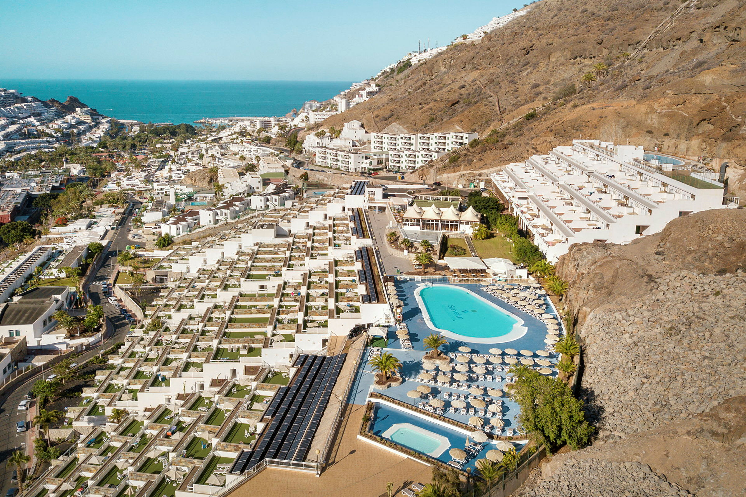 Luftfoto af et hotelområde med pools, liggestole og terrasser, beliggende ved en skråning nær kysten. (AI-genereret tekst)