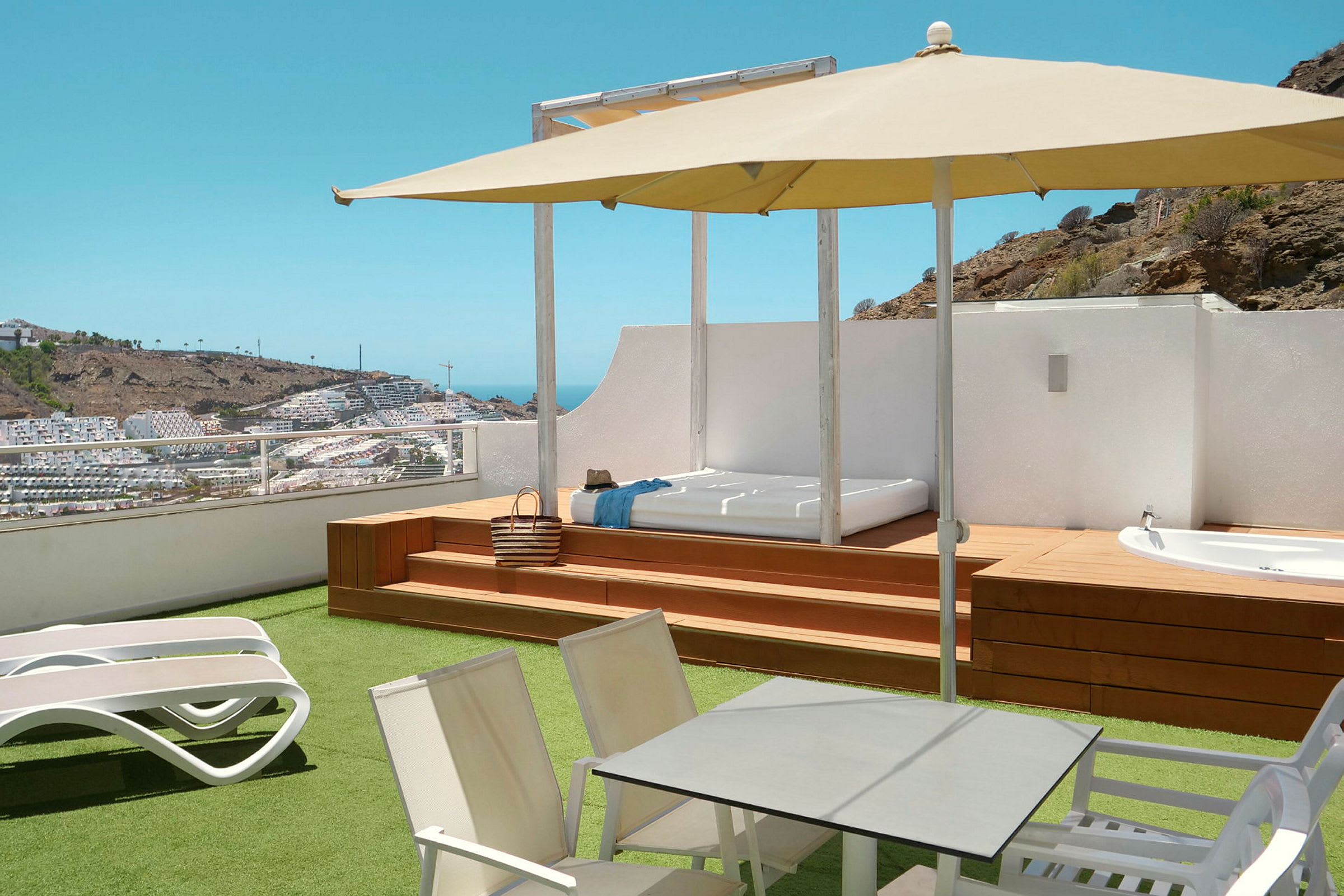Terrasse med liggestole, bord, parasol og udsigt over et kuperet område med bygninger. (AI-genereret tekst)