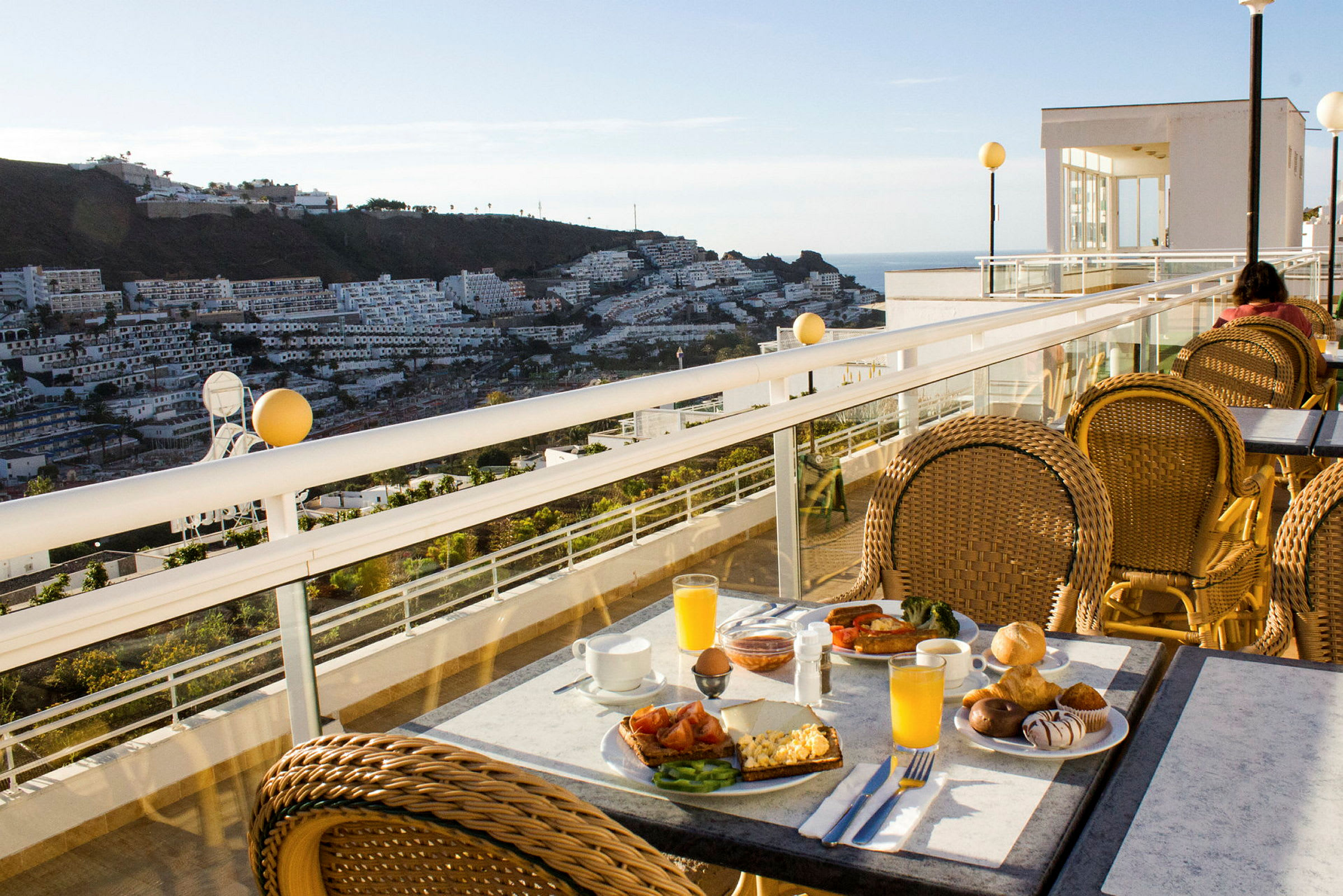 Udsigt fra en terrasse med morgenmad på et bord og boligbygninger i baggrunden. (AI-genereret tekst)