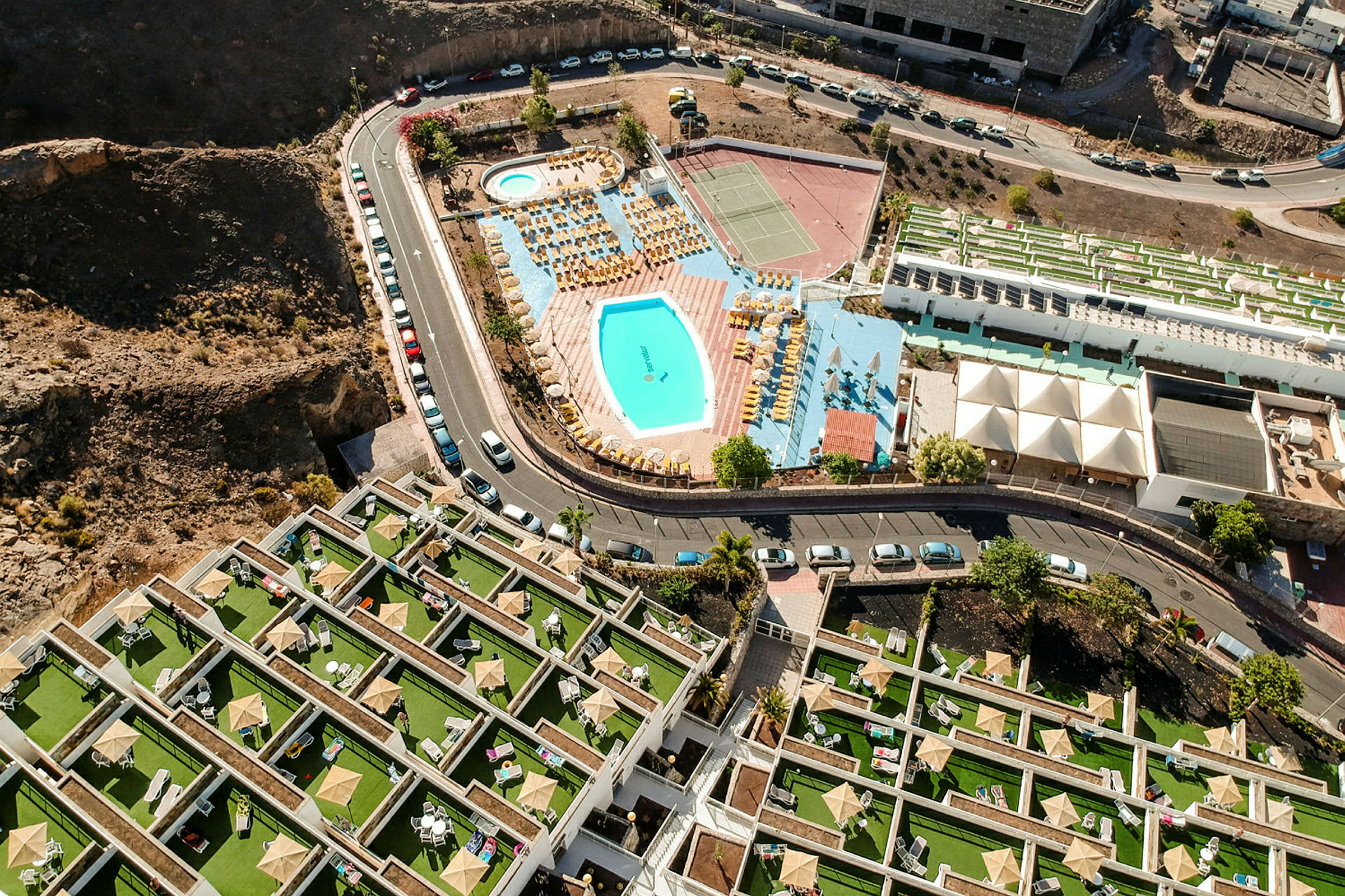 Luftfoto af hotelområde med pool, solsenge og tennisbaner. (AI-genereret tekst)