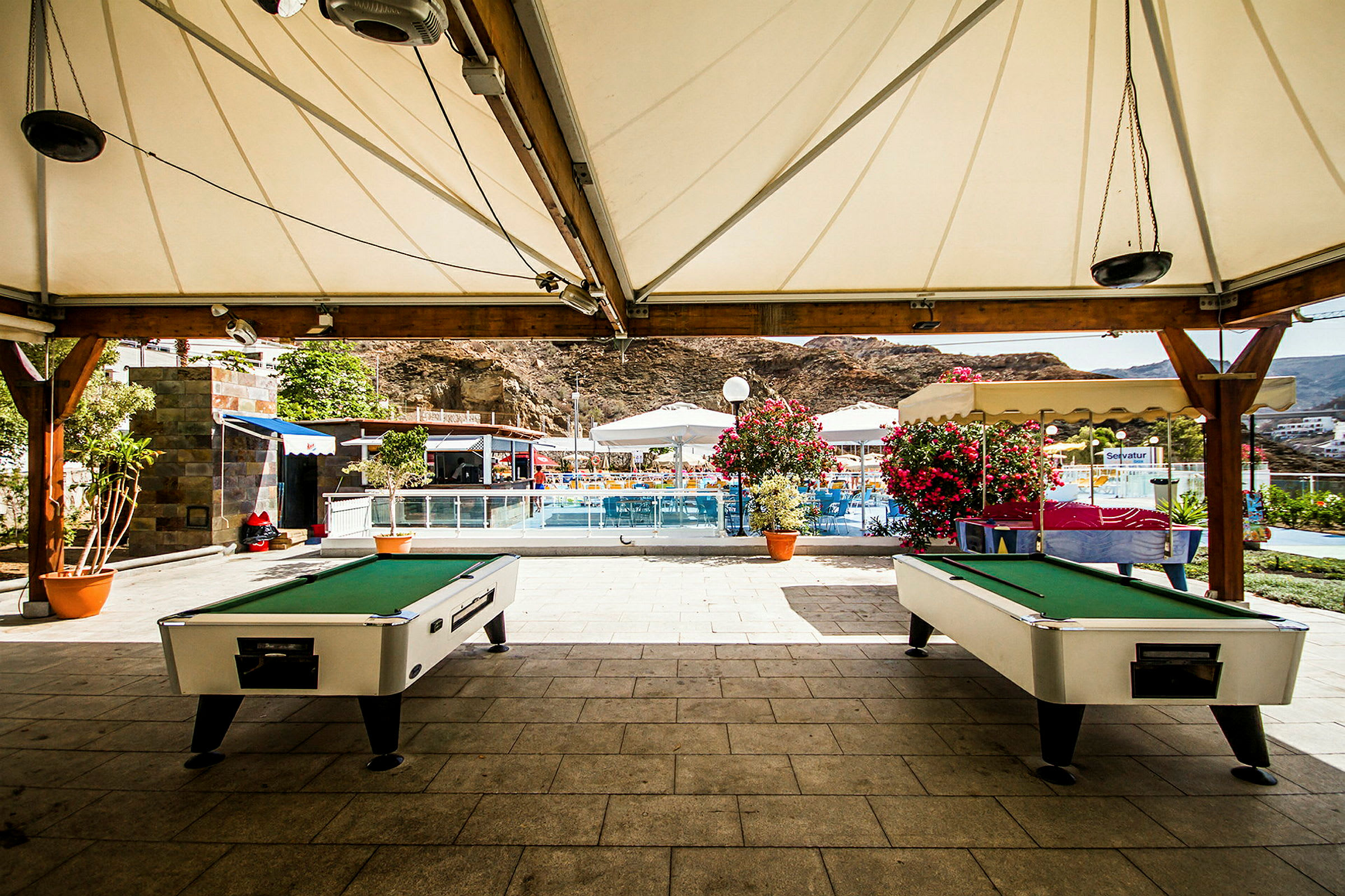 Under en teltlignende struktur er der placeret to billardborde. I baggrunden ses en pool omgivet af parasoller og blomstrende planter. (AI-genereret tekst)