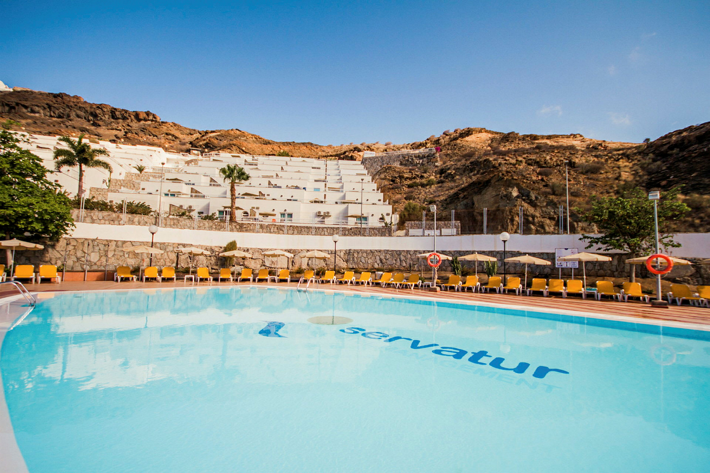Udendørs pool ved et feriehotel med liggestole og udsigt til en skråning med terrassebygninger. (AI-genereret tekst)