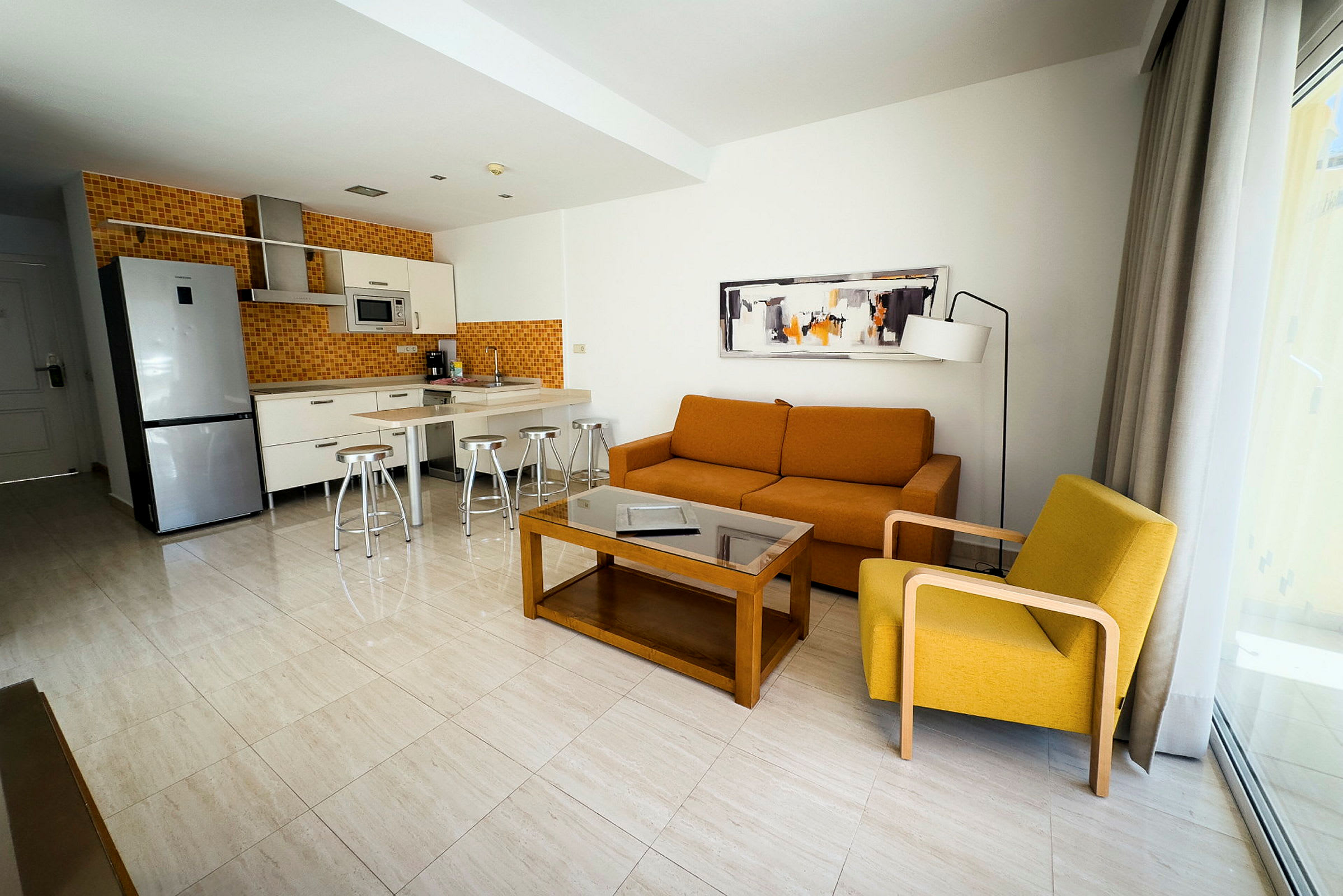 Moderne hotelværelse med køkken og stuemøbler, herunder orange sofa og gul lænestol. (AI-genereret tekst)