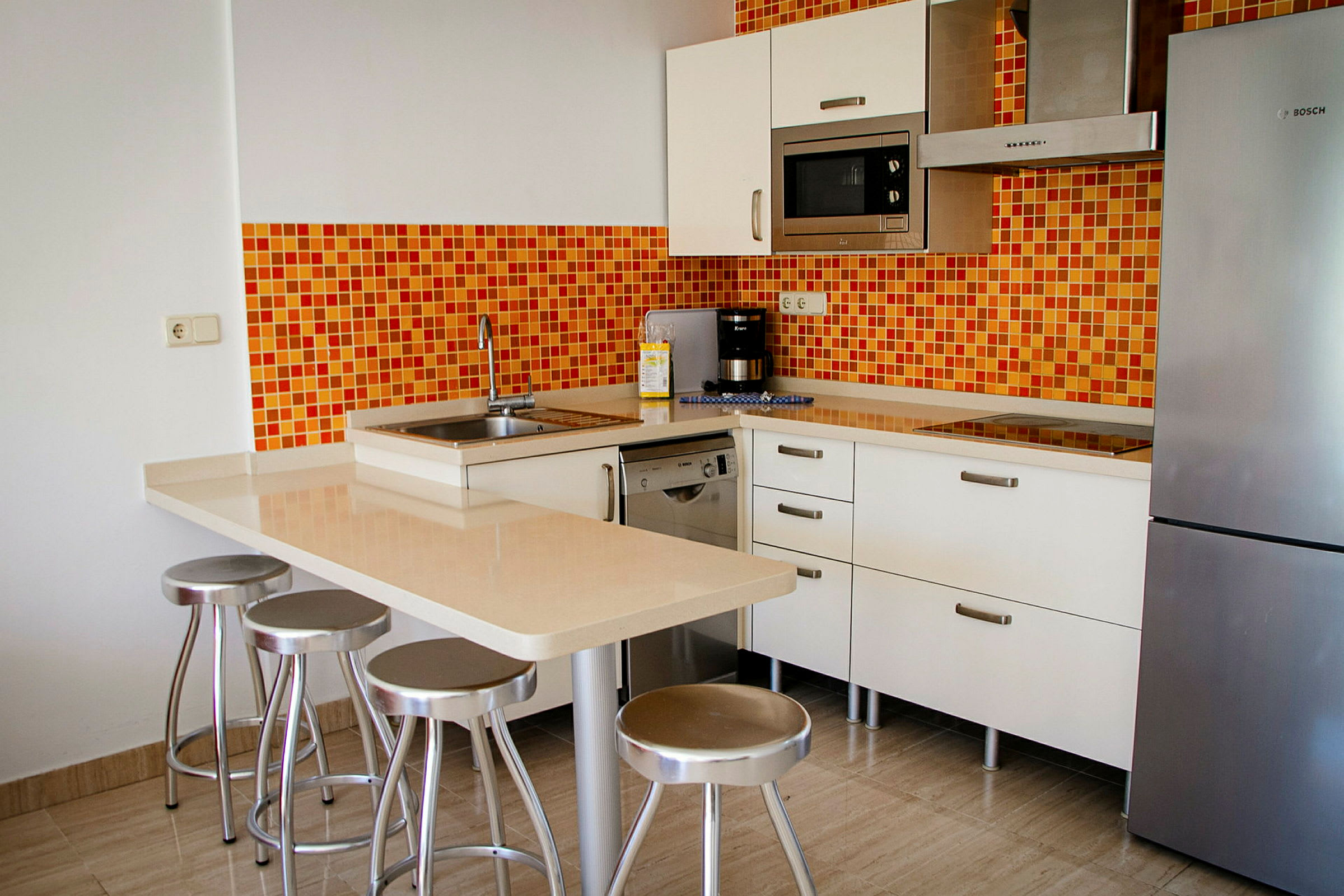 Moderne køkken med orange mosaikvæg, køleskab, mikrobølgeovn og en køkkenø med barstole. (AI-genereret tekst)
