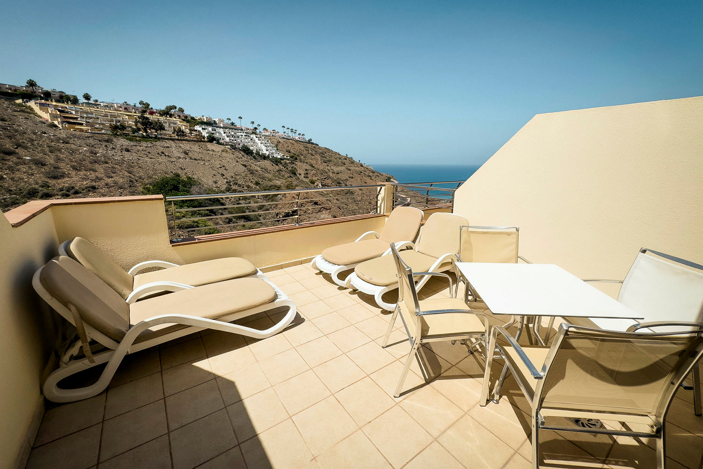 Terrasse med solsenge og bord med stole, udsigt over havet. (AI-genereret tekst)