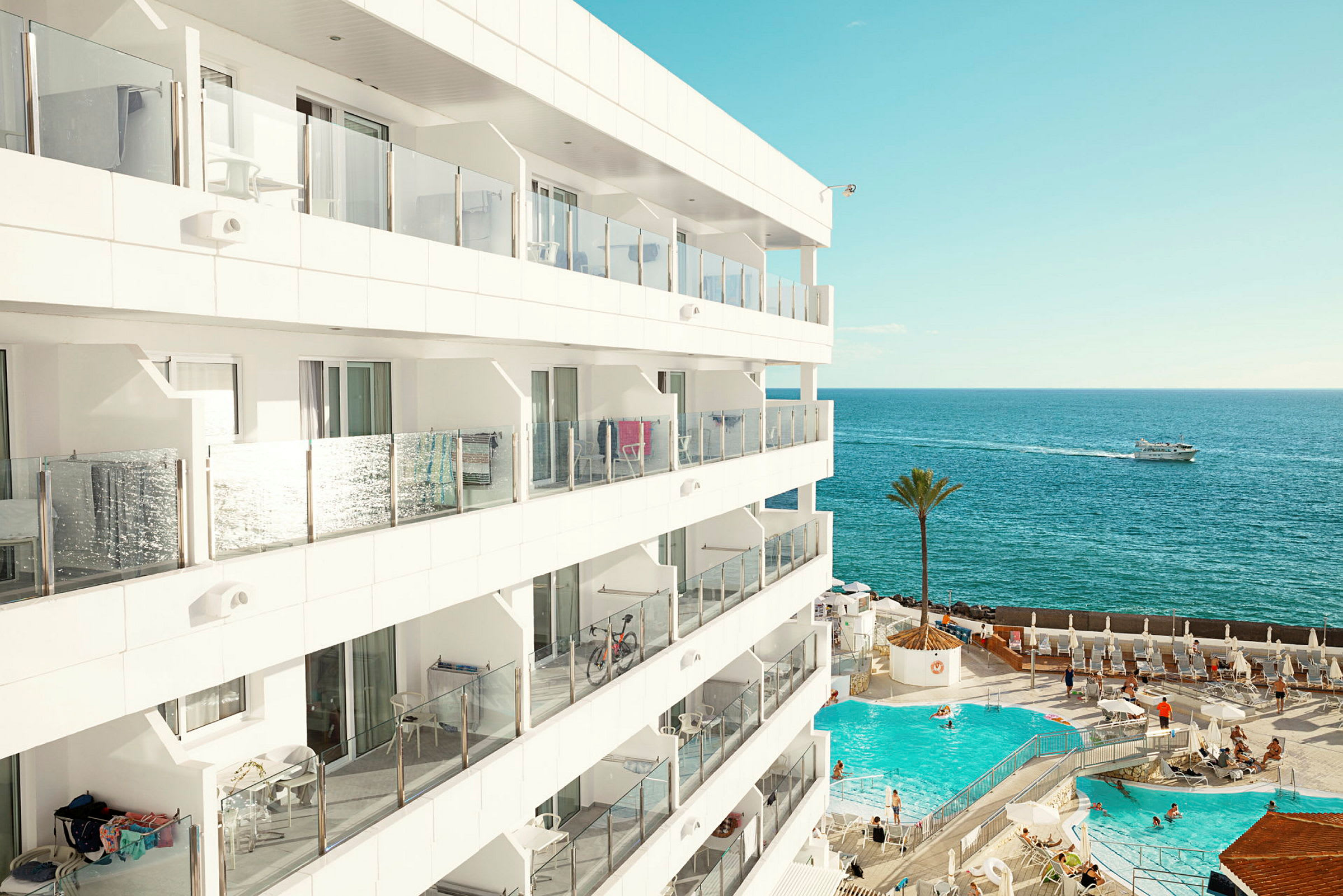 Udsigt over et moderne hotel med altaner, en udendørs pool og havet i baggrunden. (AI-genereret tekst)