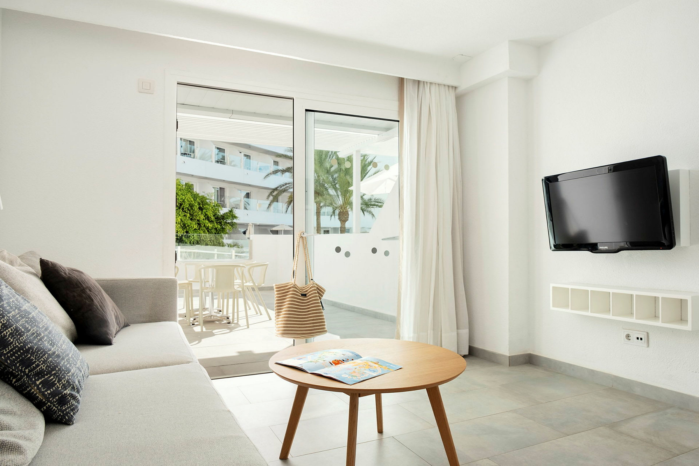 En stue med sofa, sofabord og en vægmonteret TV. Balkon med bord og stole synlig gennem glasdørene. (AI-genereret tekst)