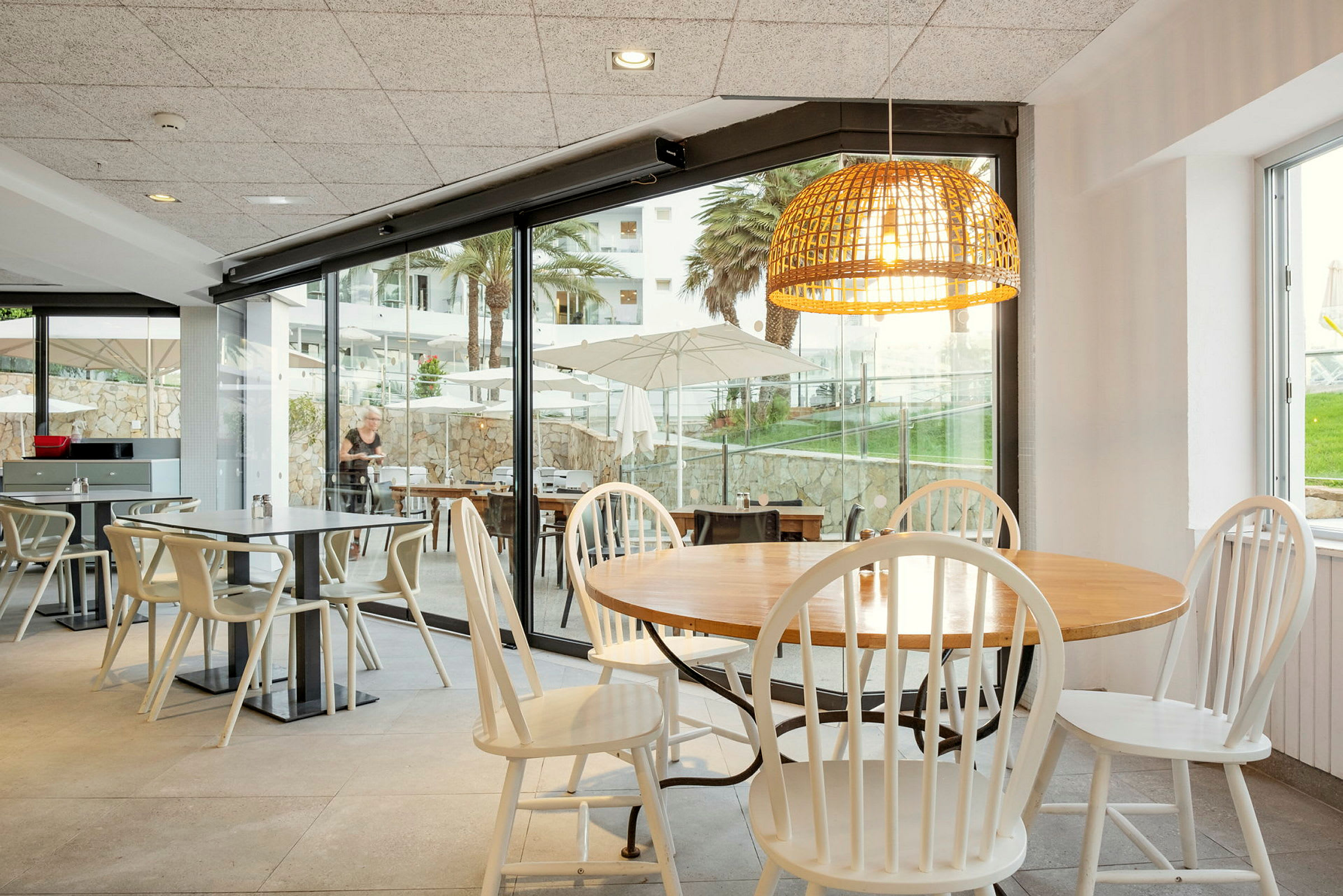 En restaurant med borde og stole. Store glasvinduer viser udsigten med parasoller, grønne områder og palmer. (AI-genereret tekst)