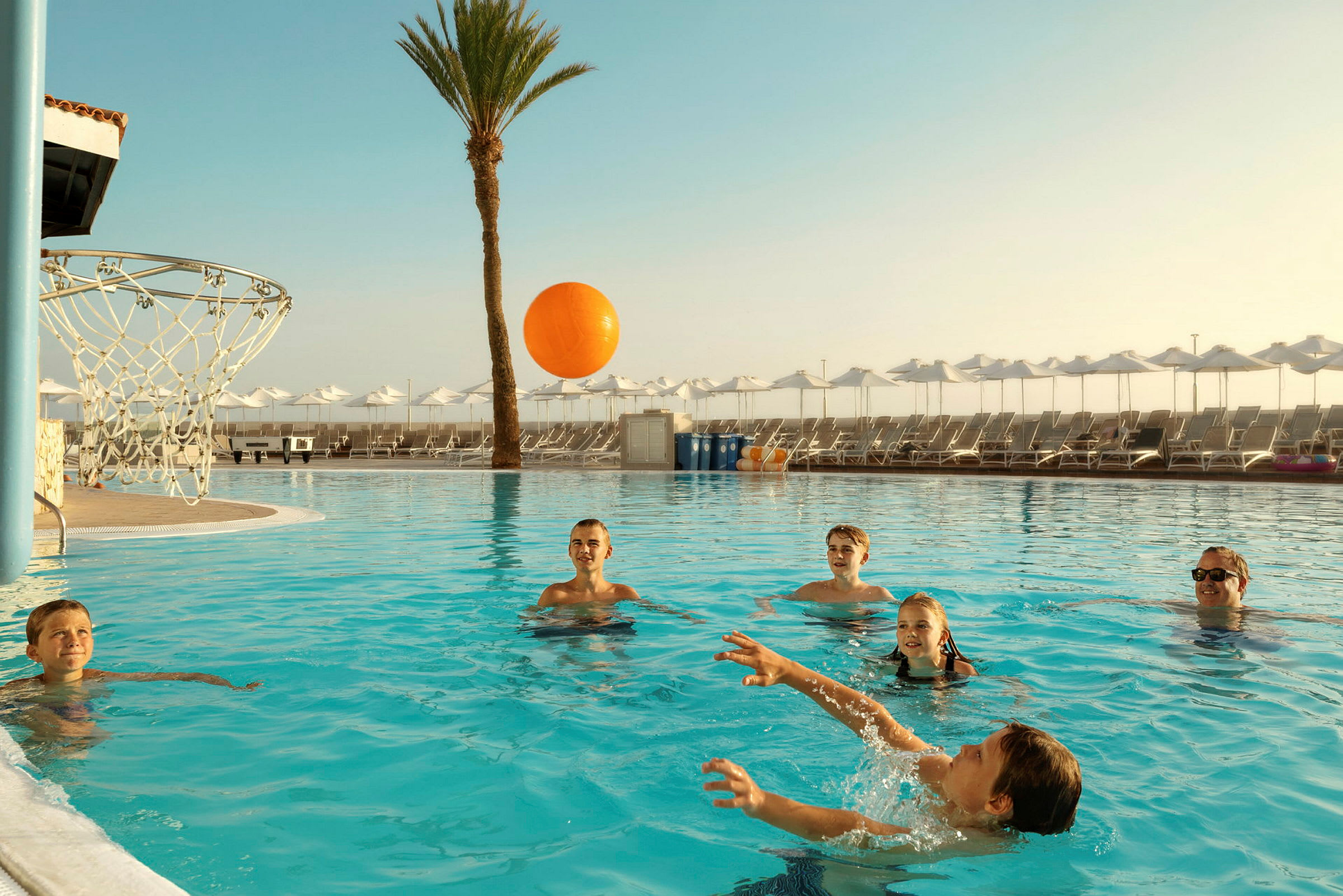Swimmingpool med en basketballkurv og personer, der leger med en orange bold. (AI-genereret tekst)
