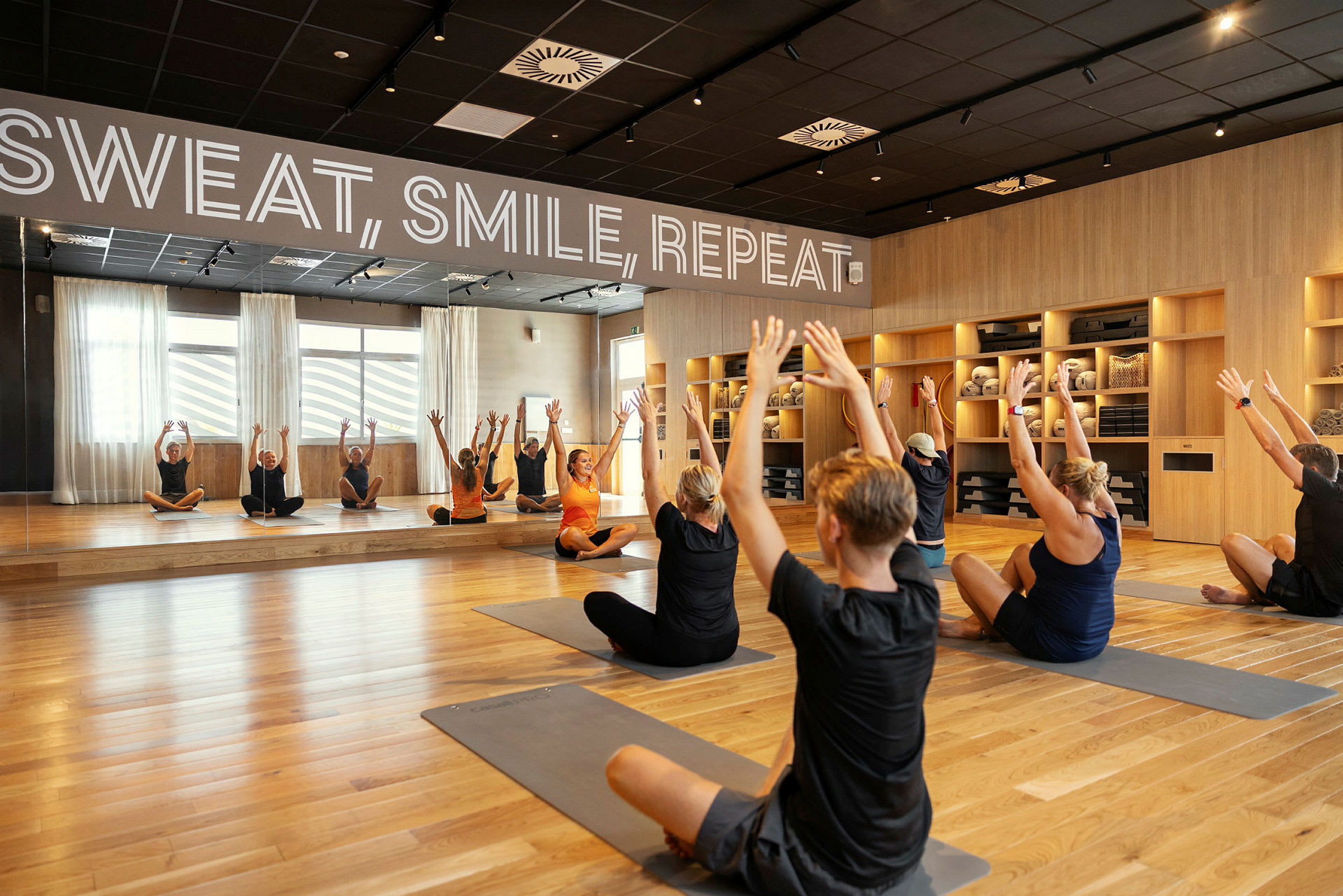 Et træningslokale med spejlvæg, hvor flere personer udfører strækøvelser på yogamåtter. (AI-genereret tekst)