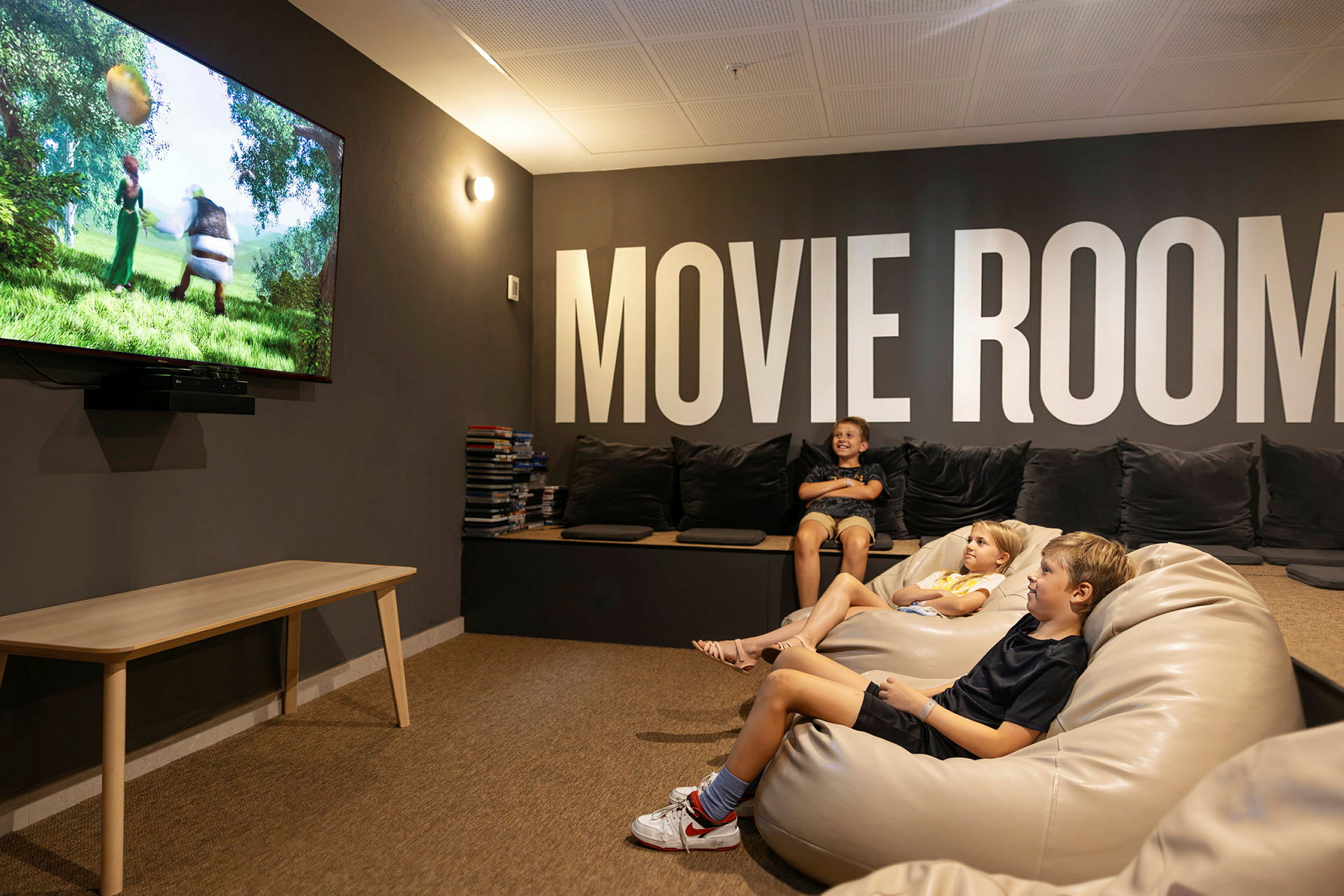 Et værelse med bløde siddepladser og et stort tv, der viser en animationsfilm. Væggene har teksten "MOVIE ROOM". (AI-genereret tekst)