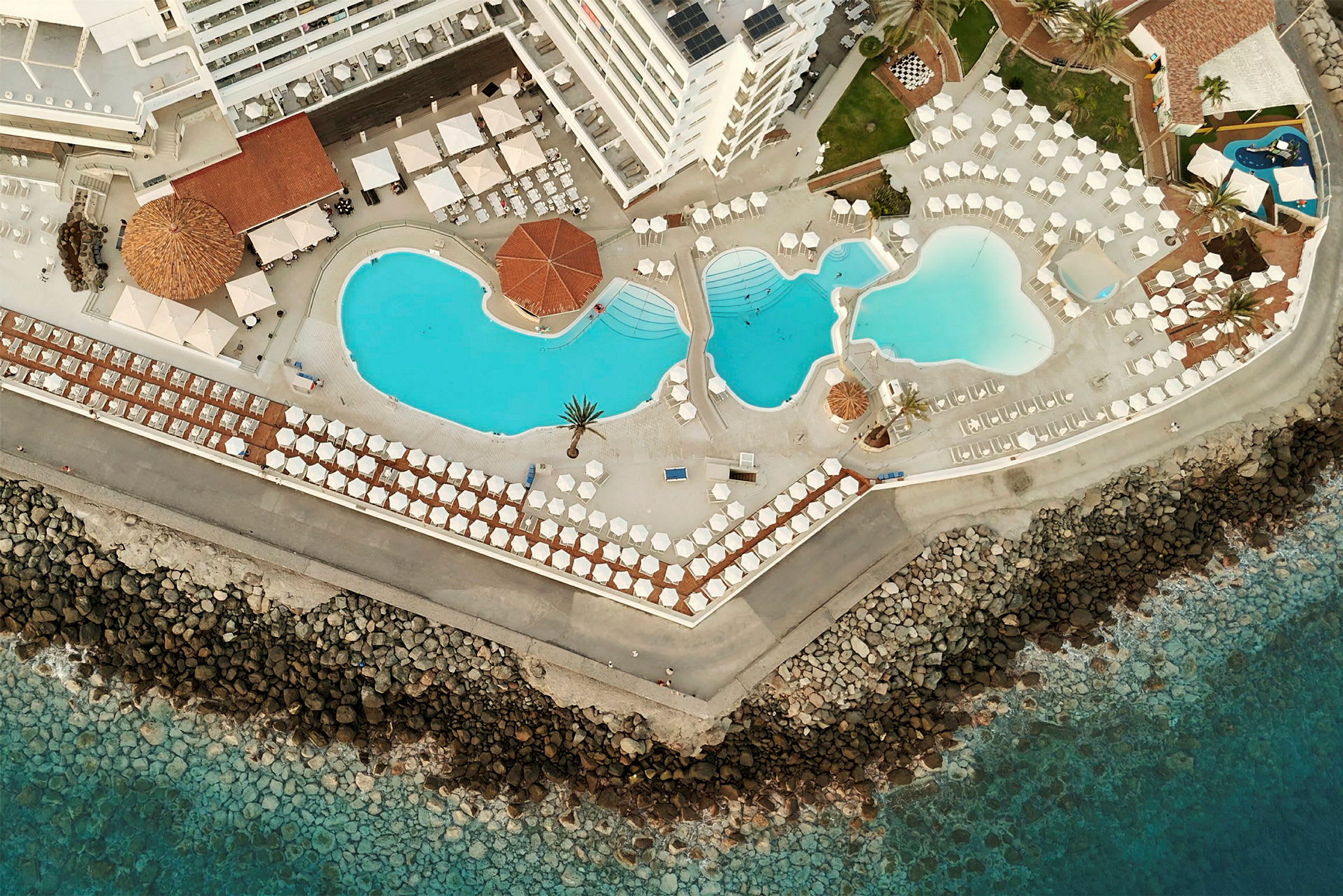 Luftfoto af et hotelområde med flere swimmingpools tæt på havet. (AI-genereret tekst)