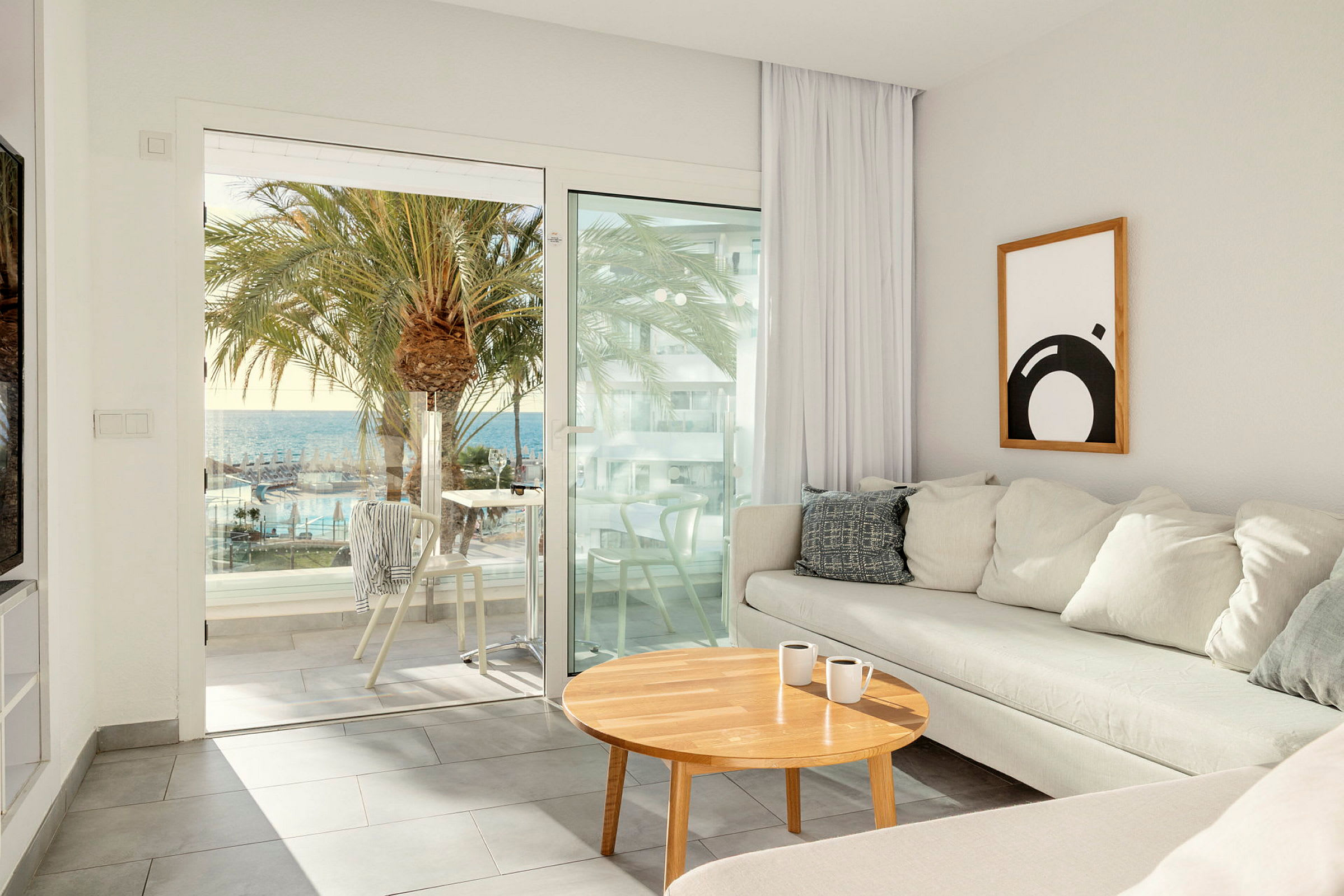En lys stue med en beige sofa og et træbord har udsigt gennem glasdøre mod en terrasse med udsigt over palmer, en pool og havet. (AI-genereret tekst)
