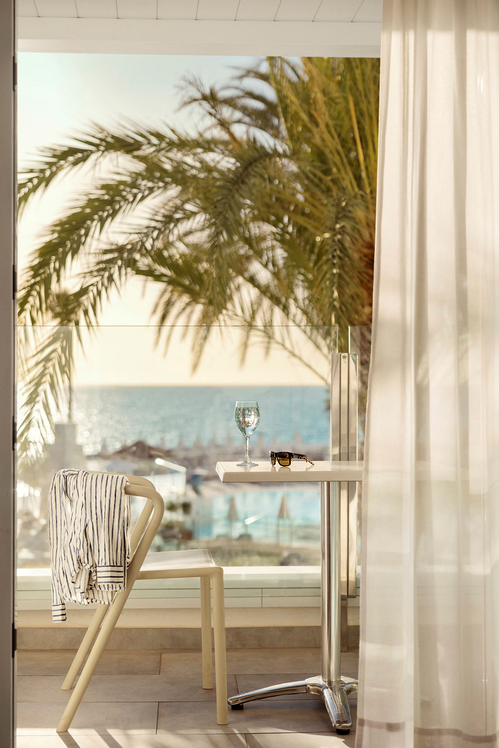 Et bord med et glas vin og et par solbriller står på en terrasse med udsigt over stranden og havet. En stol har en stribet skjorte hængende over ryggen og står ved siden af bordet. (AI-genereret tekst)