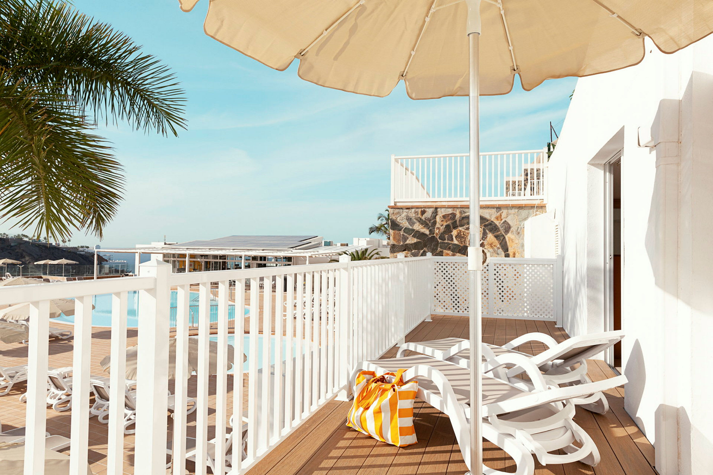 Udsigt over en terrasse med liggestole, parasol og en strandtaske. I baggrunden ses en pool og palmer. (AI-genereret tekst)