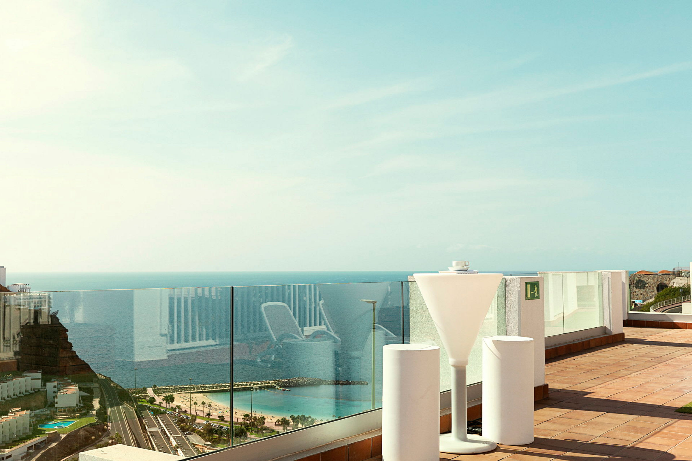 Udsigt fra en terrasse med glasrækværk, der viser havet og en strand. (AI-genereret tekst)