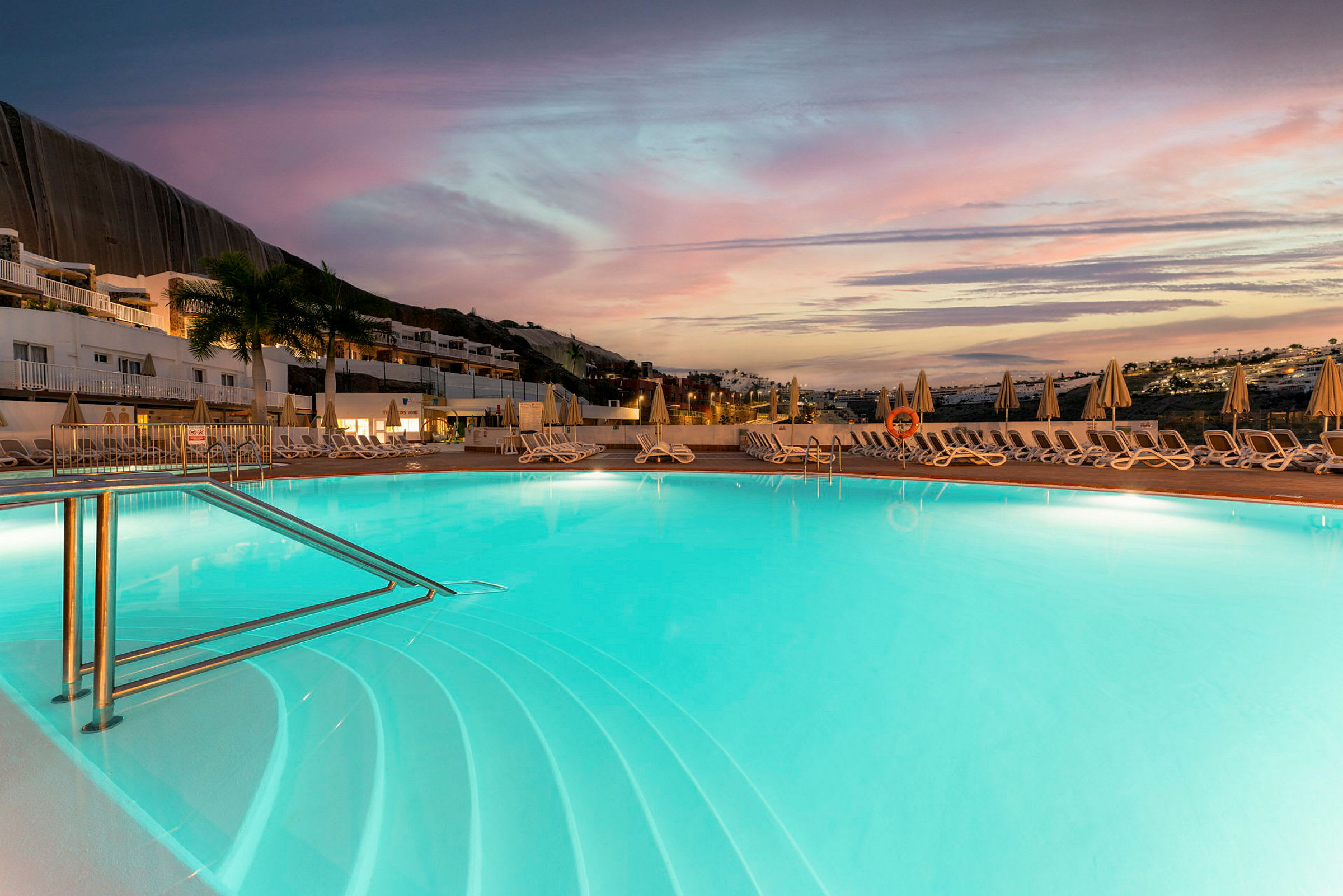 Udendørs swimmingpool ved solnedgang omgivet af liggestole og parasoller. (AI-genereret tekst)