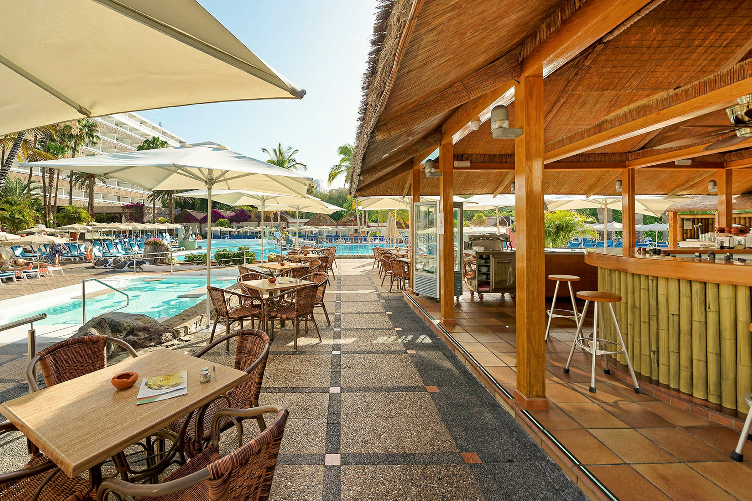 En poolbar med udendørs servering og borde, stole samt parasoller ved et poolområde med liggestole og planter. (AI-genereret tekst)
