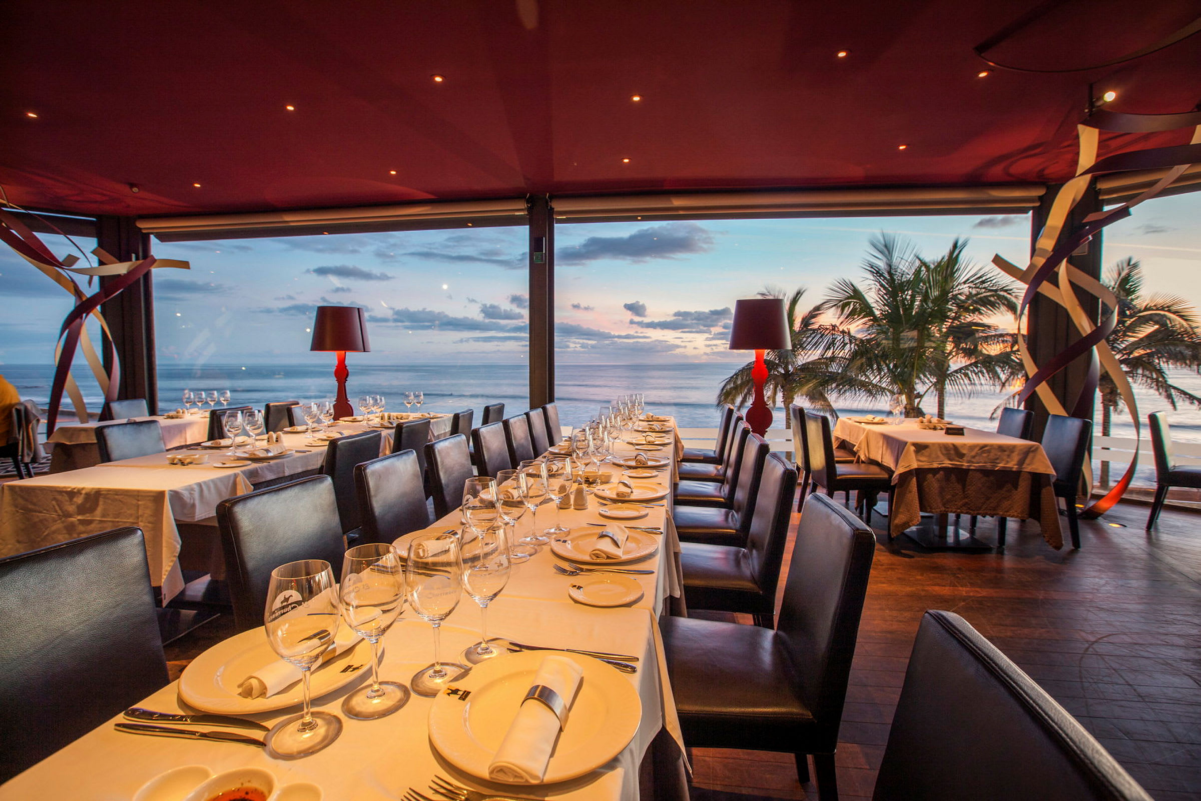 En restaurant med dækkede borde og udsigt over havet ved solnedgang. (AI-genereret tekst)