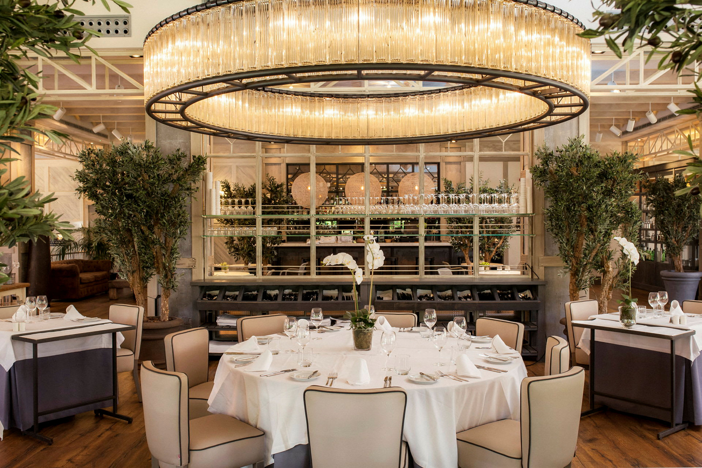 Elegant restaurantinteriør med en rund lampe i loftet over et dækket bord omgivet af stole. Rummet har også store planter og en moderne indretning. (AI-genereret tekst)