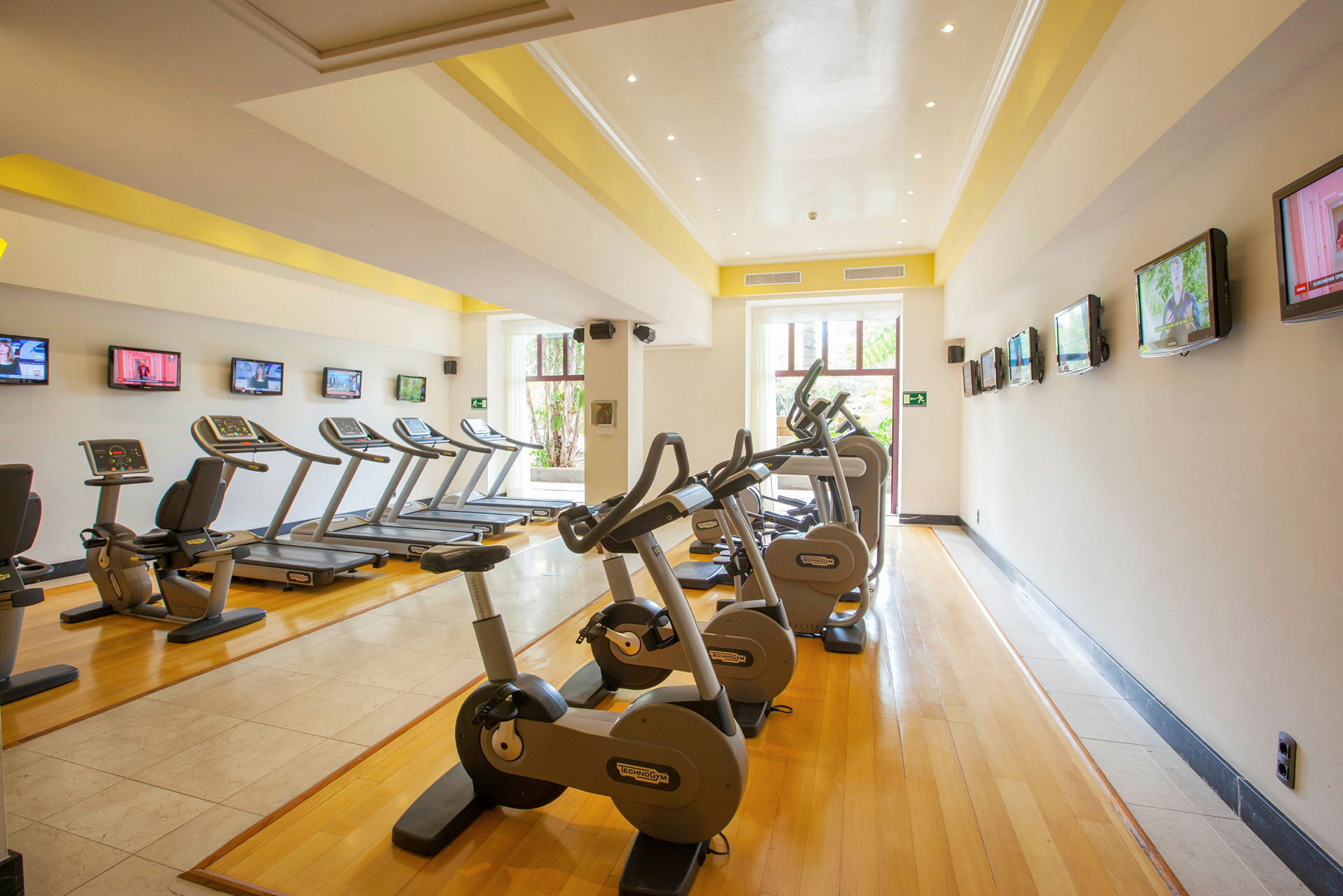 Billede af et fitnesscenter med motionscykler og løbebånd på række langs et rum med trægulv og flere tv-skærme på væggene. (AI-genereret tekst)