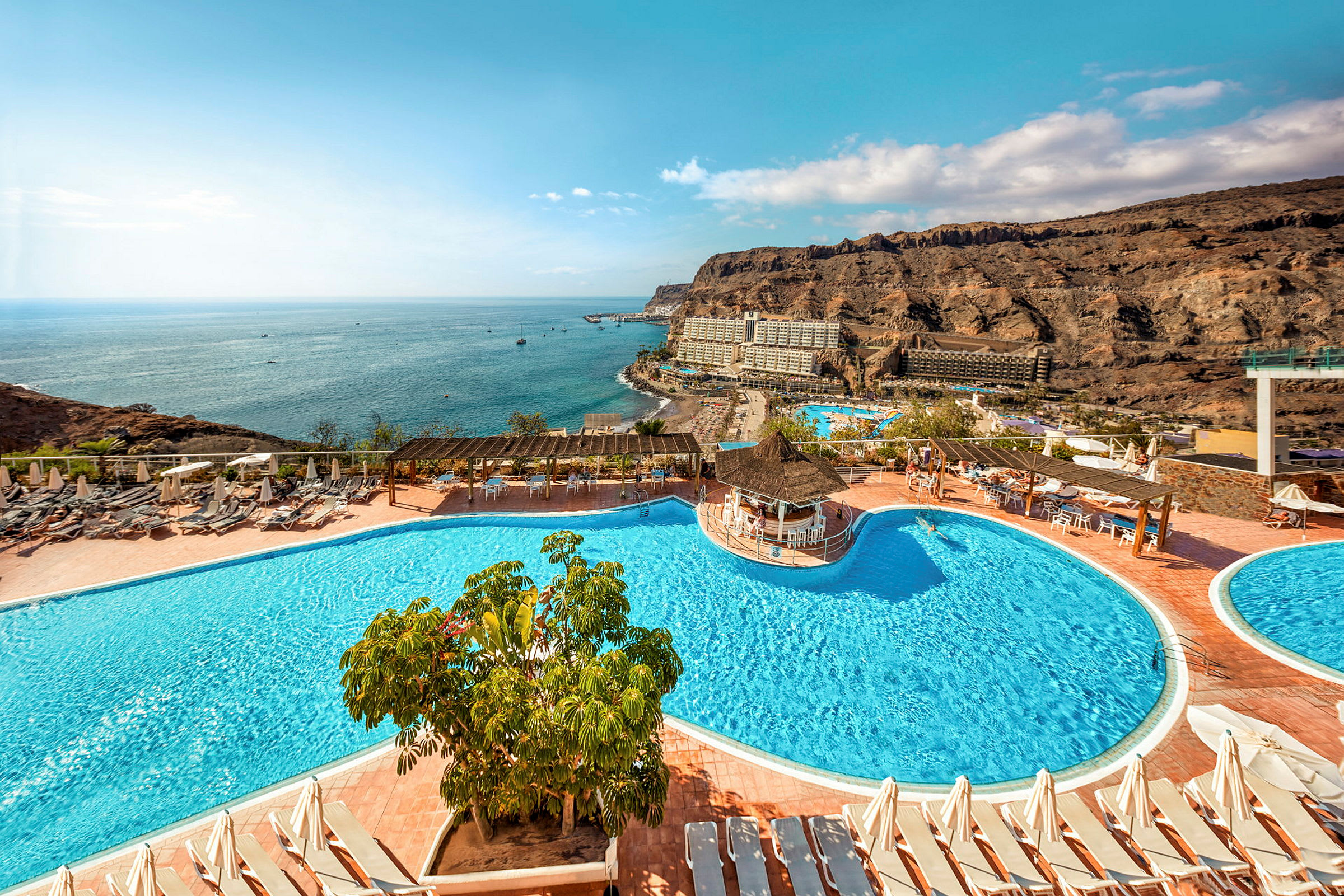 Hotelområde med swimmingpools, liggestole og udsigt over havet samt de omkringliggende klipper. (AI-genereret tekst)
