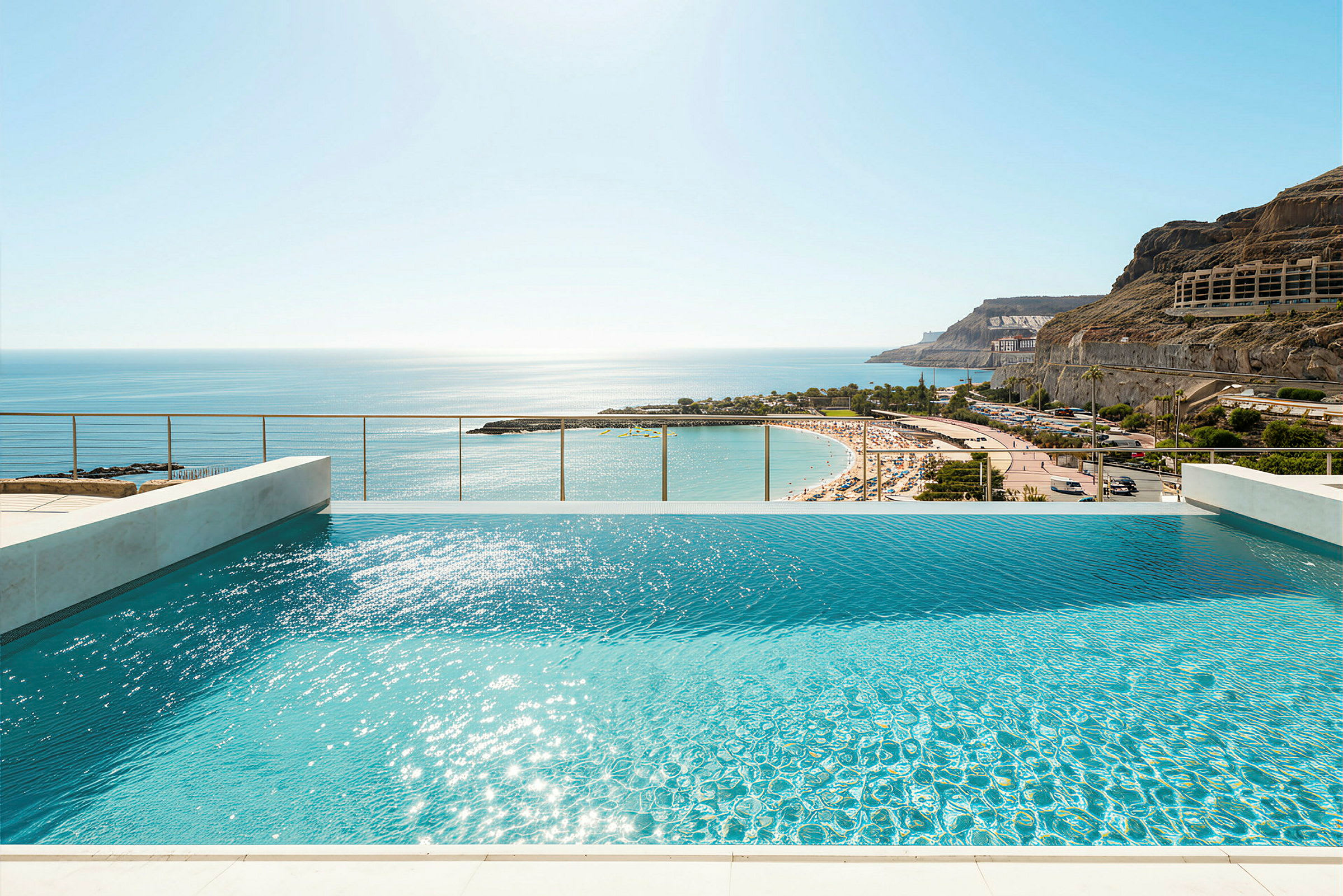 Udsigt over en infinitypool med solskin på vandoverfladen og en sandstrand i baggrunden. (AI-genereret tekst)