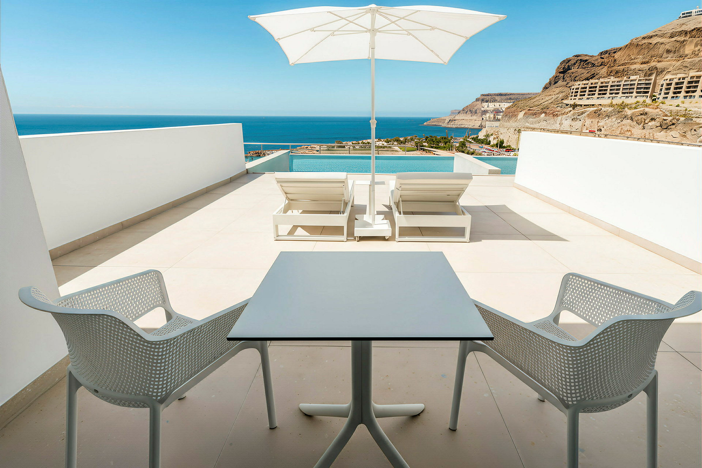 En terrasse med to liggestole, parasol og en lille siddegruppe med udsigt over havet. (AI-genereret tekst)