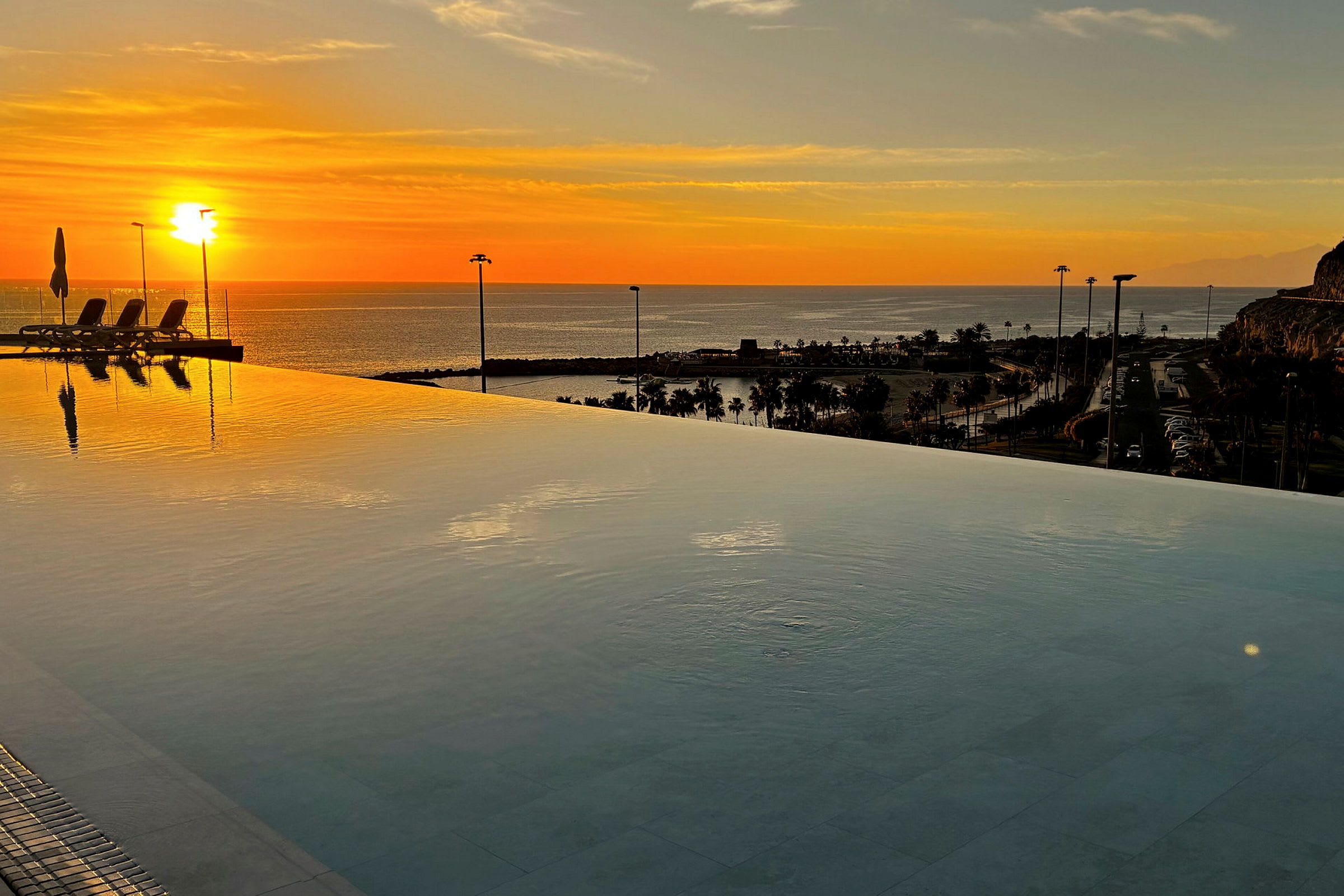 En infinitypool med udsigt over havet ved solnedgang. Solen spejler sig i vandet, og palmer anes i baggrunden. (AI-genereret tekst)