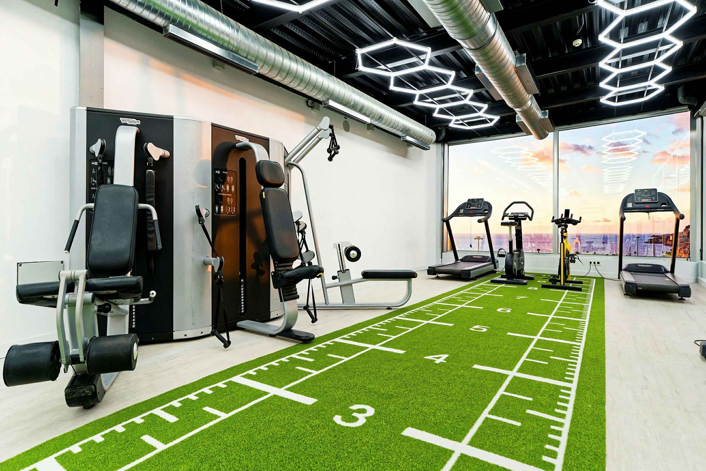Et fitnesscenter med træningsmaskiner, løbebånd og grønt gulv med markeringer. Store vinduer viser udsigt over havet og en solnedgang. (AI-genereret tekst)