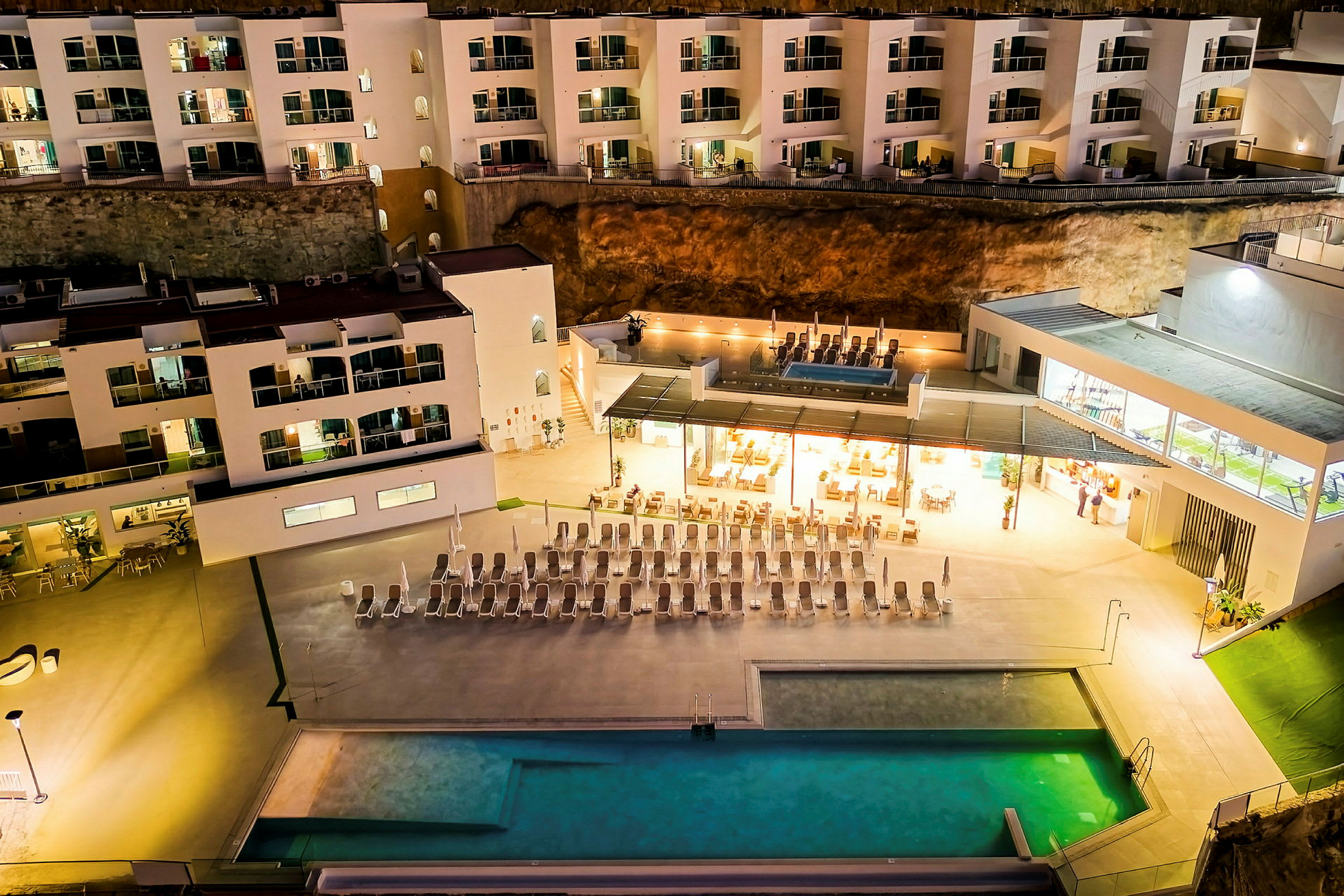 En bygning om aftenen med integreret pool og liggestole samt et oplyst restaurantområde. (AI-genereret tekst)