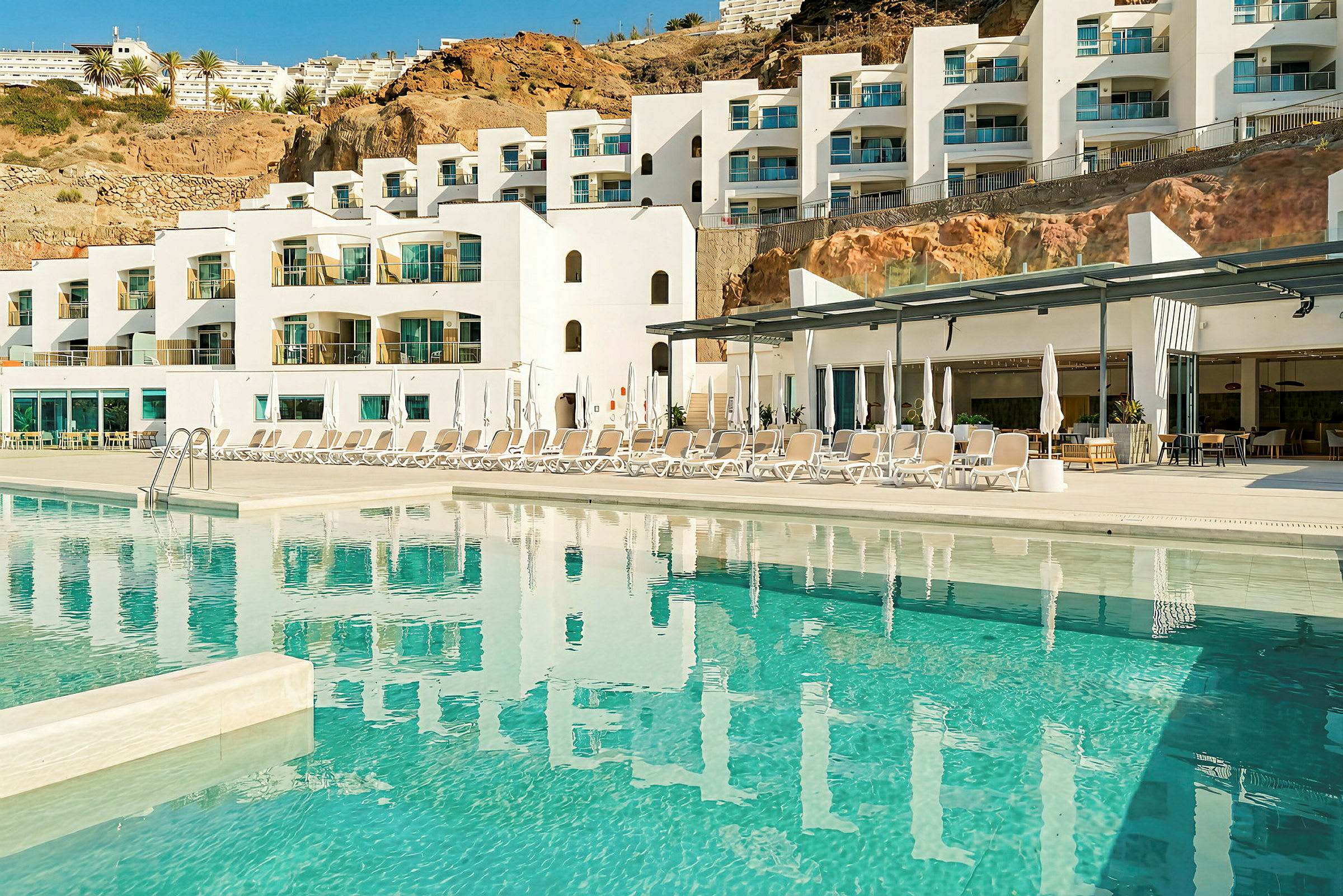 Et moderne hotel med hvid facade og flere altaner. Foran hotellet er der en pool og liggestole. (AI-genereret tekst)
