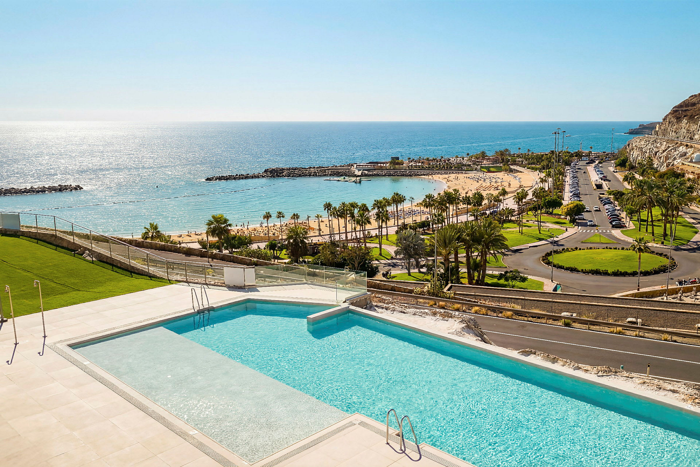 En swimmingpool med udsigt over en sandstrand, palmer og havet under en klar blå himmel. (AI-genereret tekst)