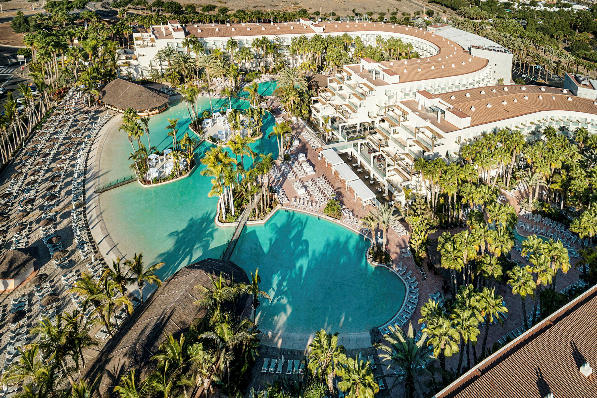 Luftfoto af en resort med store pools, palmer og solsenge. (AI-genereret tekst)