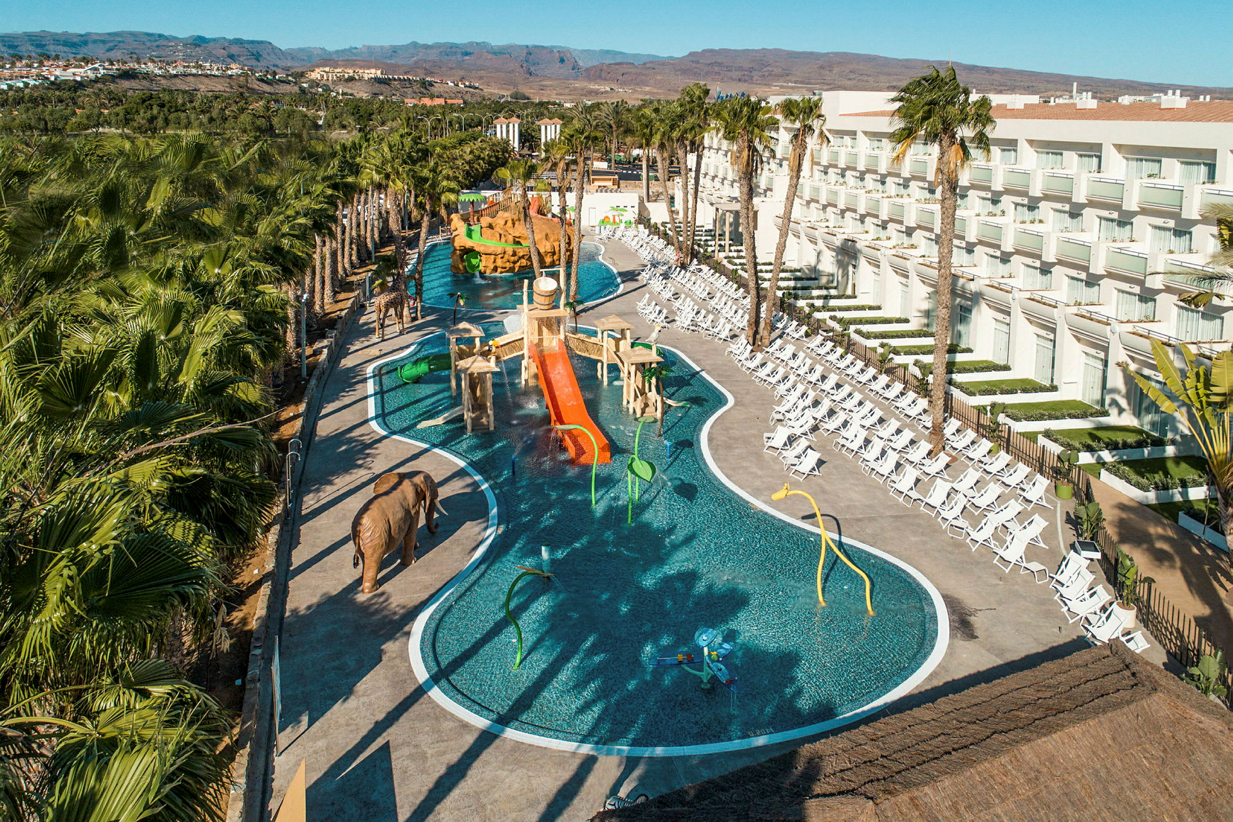Et billede af en udendørs pool ved et hotel med solsenge og palmetræer samt legefaciliteter til børn. (AI-genereret tekst)