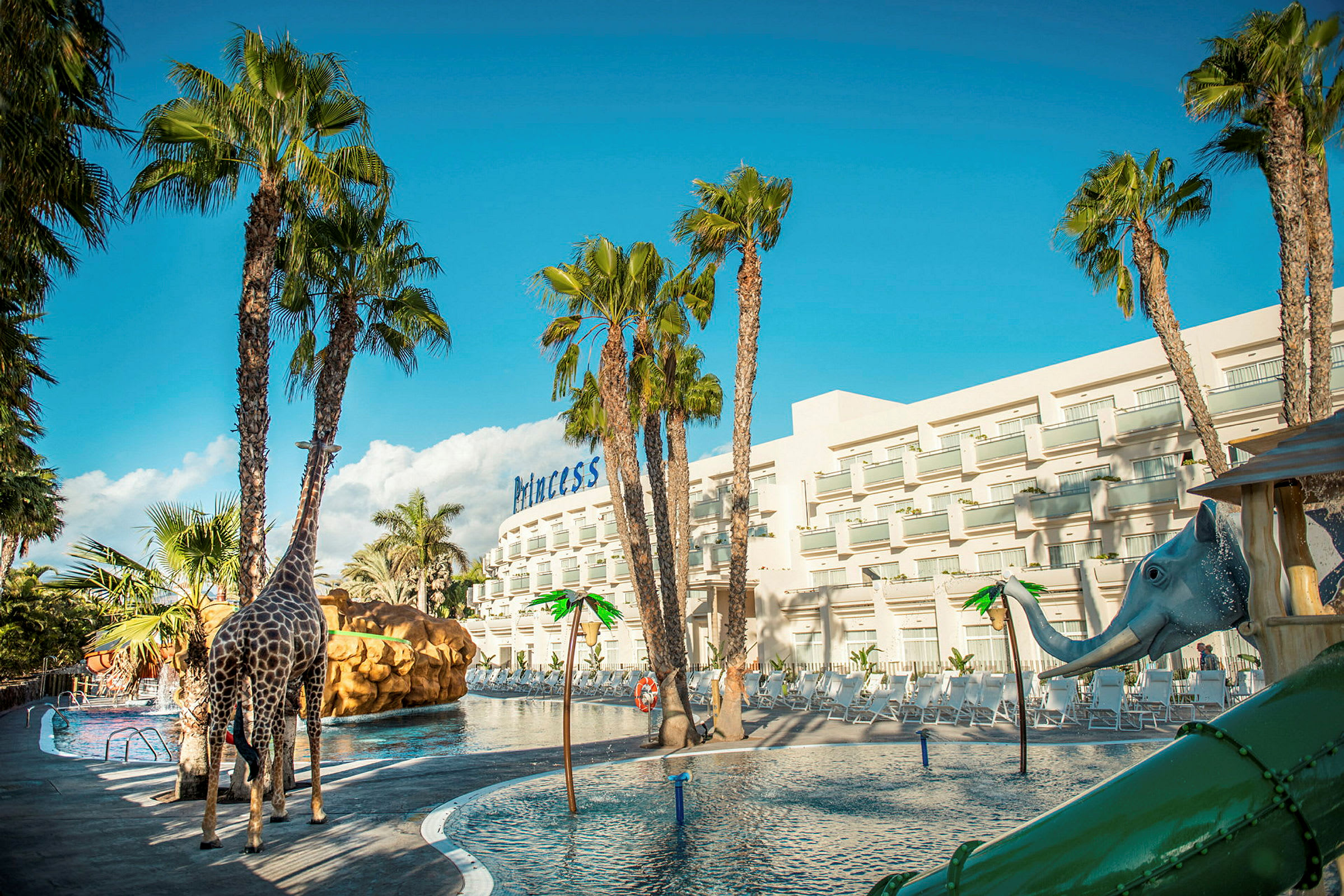 En pool med vandrutsjebane og legetøj formet som en giraf og en elefant, omgivet af palmer og en hotelbygning i baggrunden. (AI-genereret tekst)