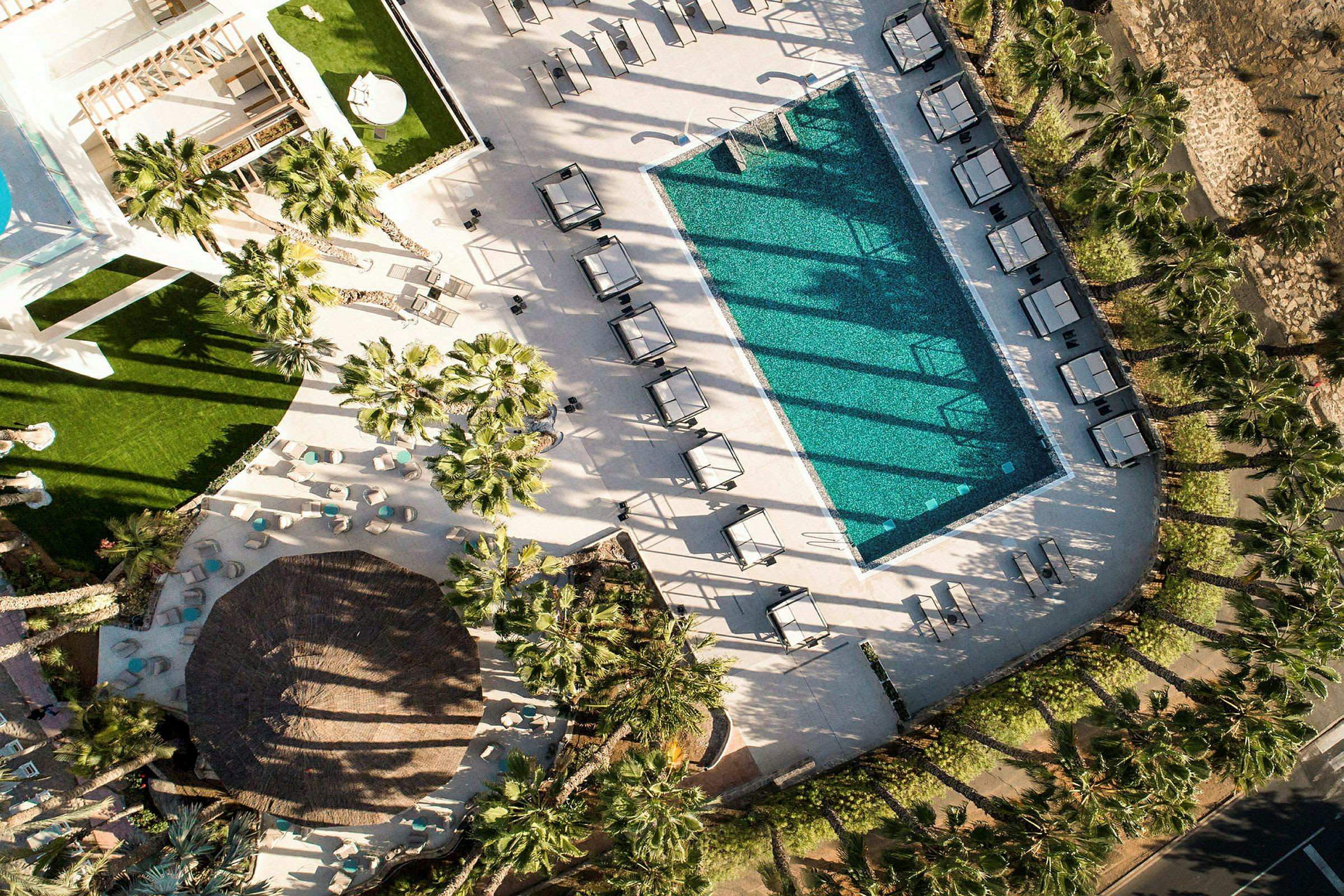 Luftfoto af en swimmingpool med liggestole og parasoller, omgivet af palmer og grønne områder. (AI-genereret tekst)