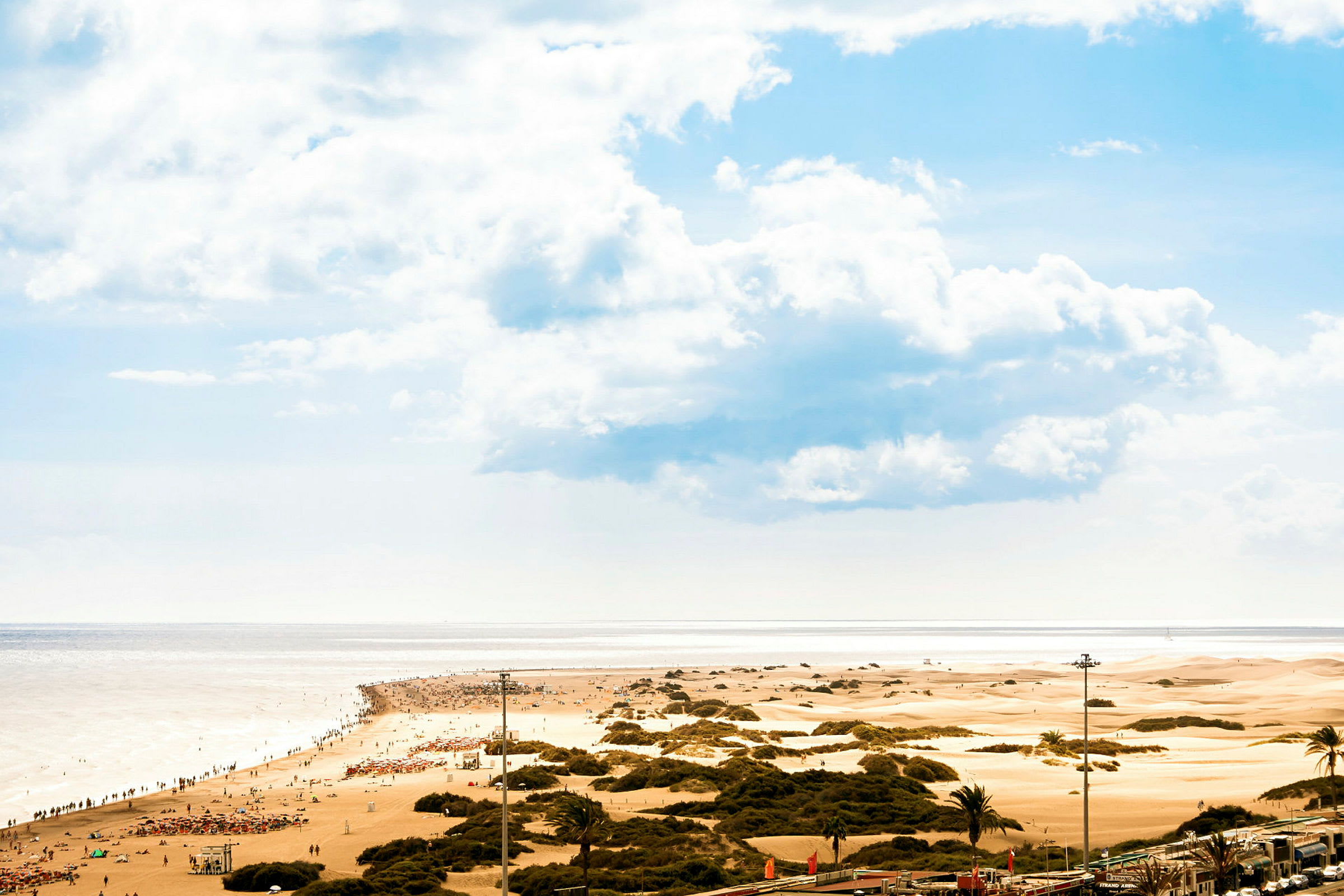 En sandstrand med mennesker samt sandklitter i baggrunden under en blå himmel. (AI-genereret tekst)