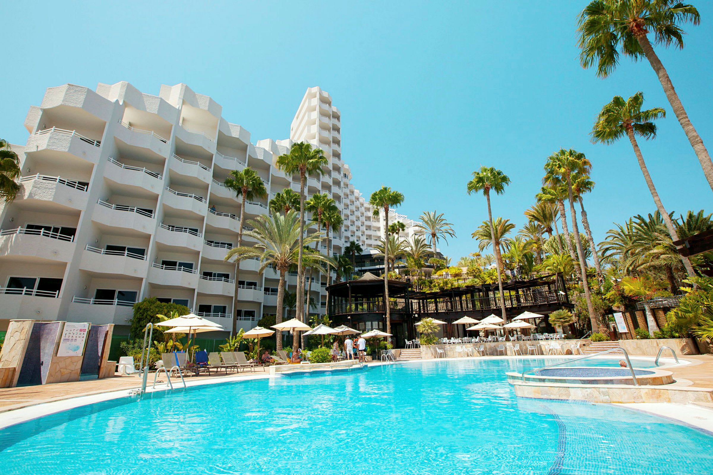 Hotelbygning med hvid facade og en stor udendørs pool omgivet af liggestole og parasoller. (AI-genereret tekst)