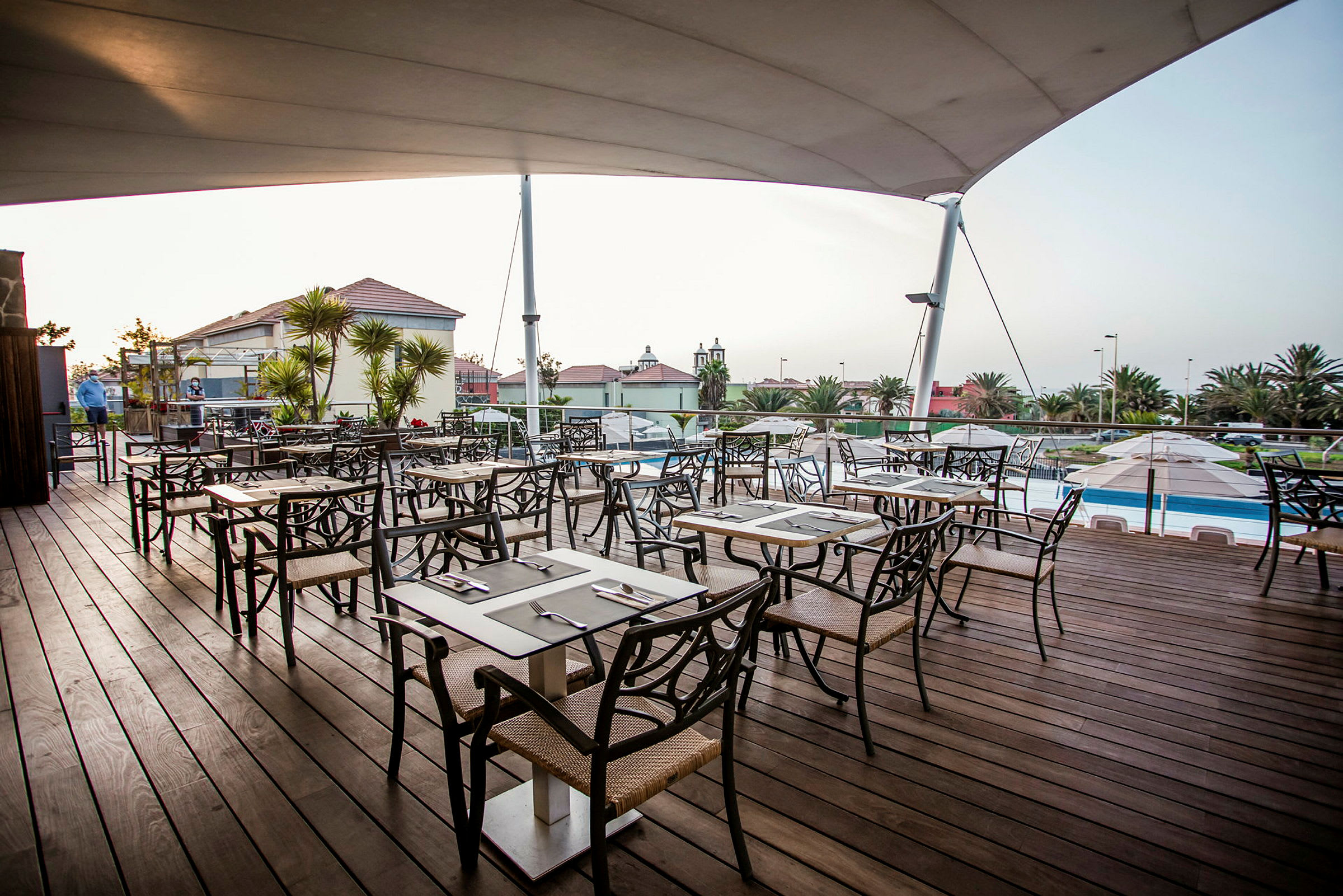 En udendørsrestaurant med træterrasse og stole omkring bordene, omgivet af grønne områder og bygninger. (AI-genereret tekst)