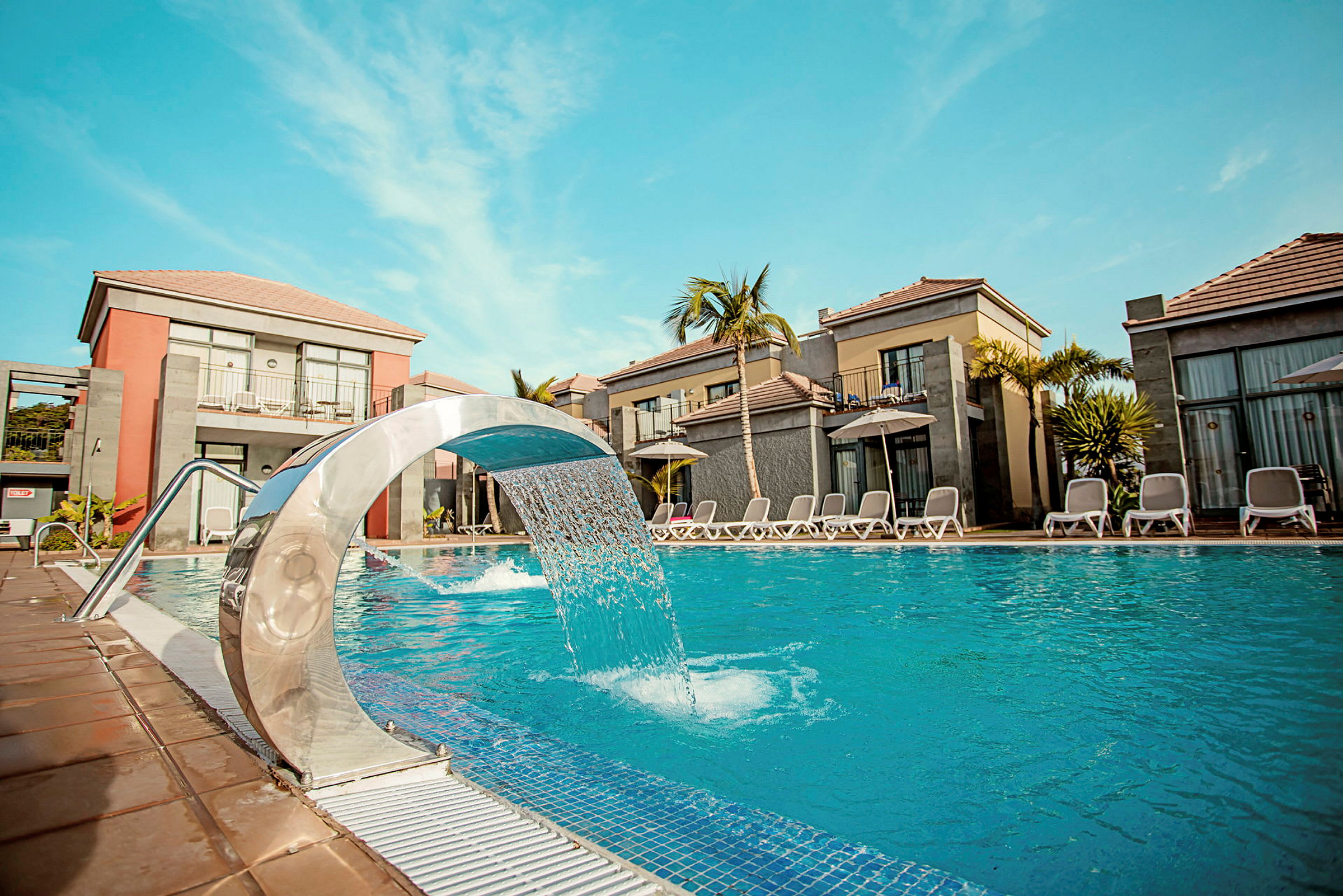 En swimmingpool med en moderne vandfontæne omgivet af solsenge og ferieboliger under en klar blå himmel. (AI-genereret tekst)