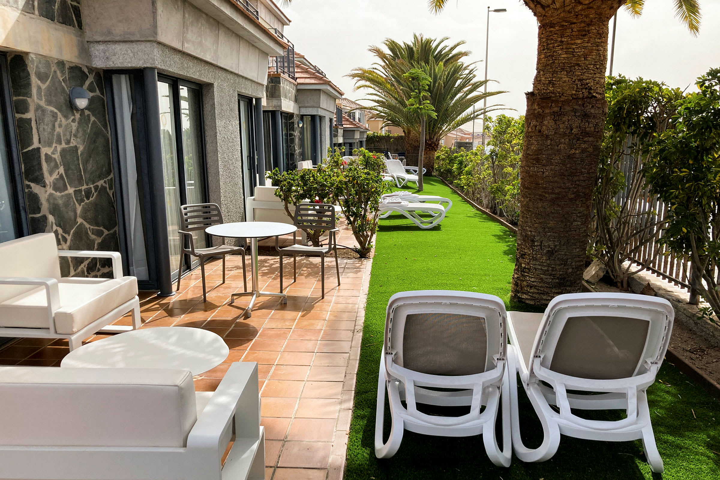 En udendørs terrasse med møbler og liggestole ved en græsplæne og træer. (AI-genereret tekst)