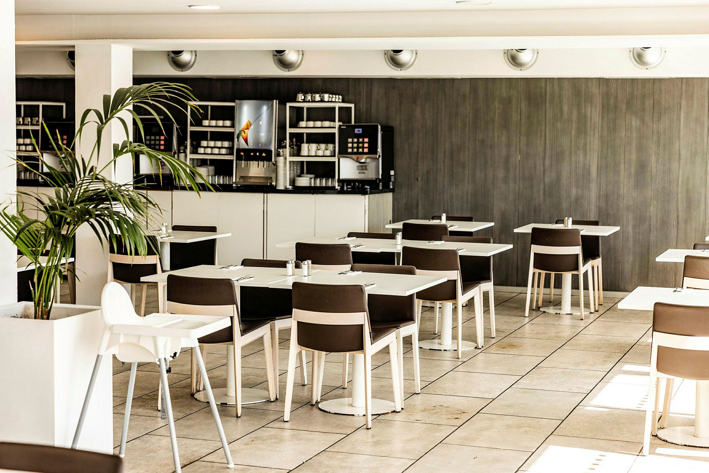 En moderne café eller restaurant med hvide borde, brune stole og en digital drikkeautomat synlig i hjørnet. Gulvet er flisebelagt, og en grøn plante står ved siden af. (AI-genereret tekst)