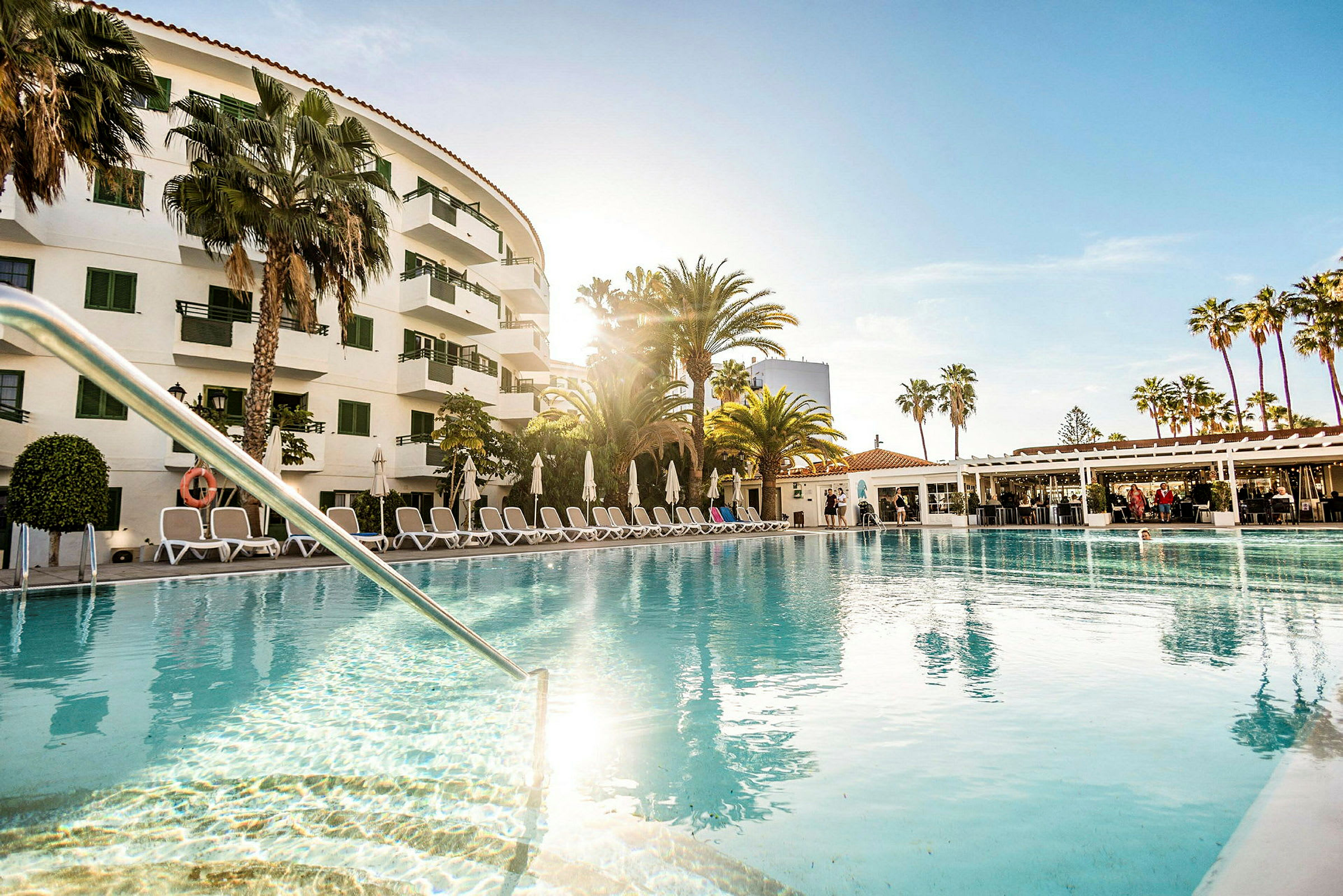 Et hotel med en udendørs pool omgivet af liggestole, parasoller og palmer. (AI-genereret tekst)
