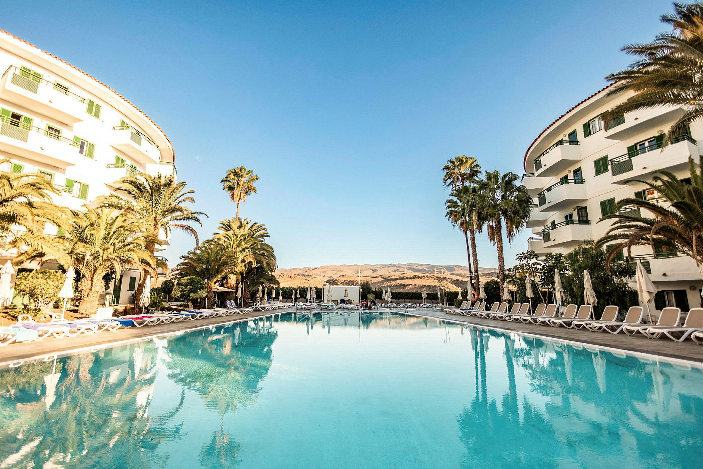 Et hotelområde med pool i forgrunden, omgivet af liggestole og parasoller samt omgivende bygninger på siderne. (AI-genereret tekst)