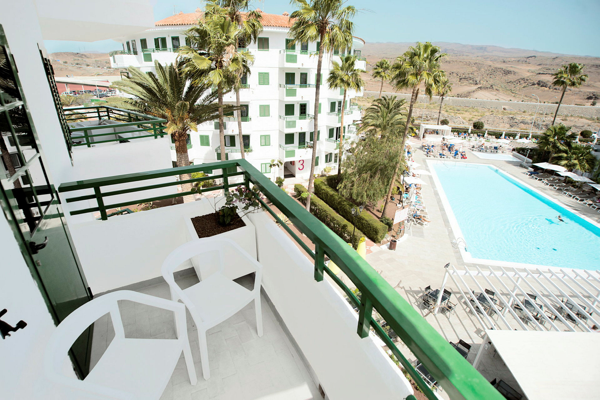 Hoteludsigt fra altan, med poolområde, solvogne, frodige palmer og lyse bygninger. (AI-genereret tekst)